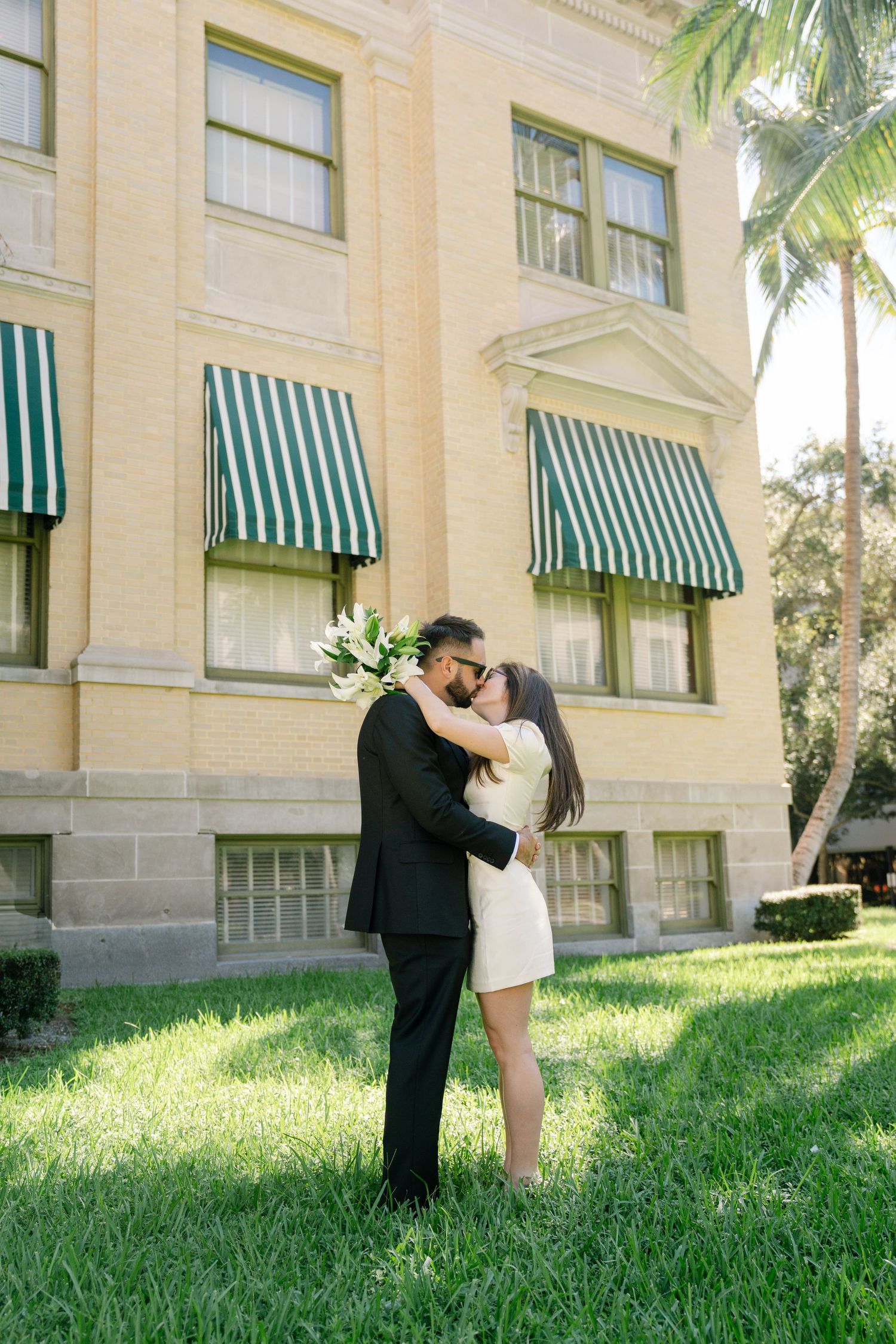 A romantic couple embraces and kisses in front of a grand building with green striped awnings and palm trees.