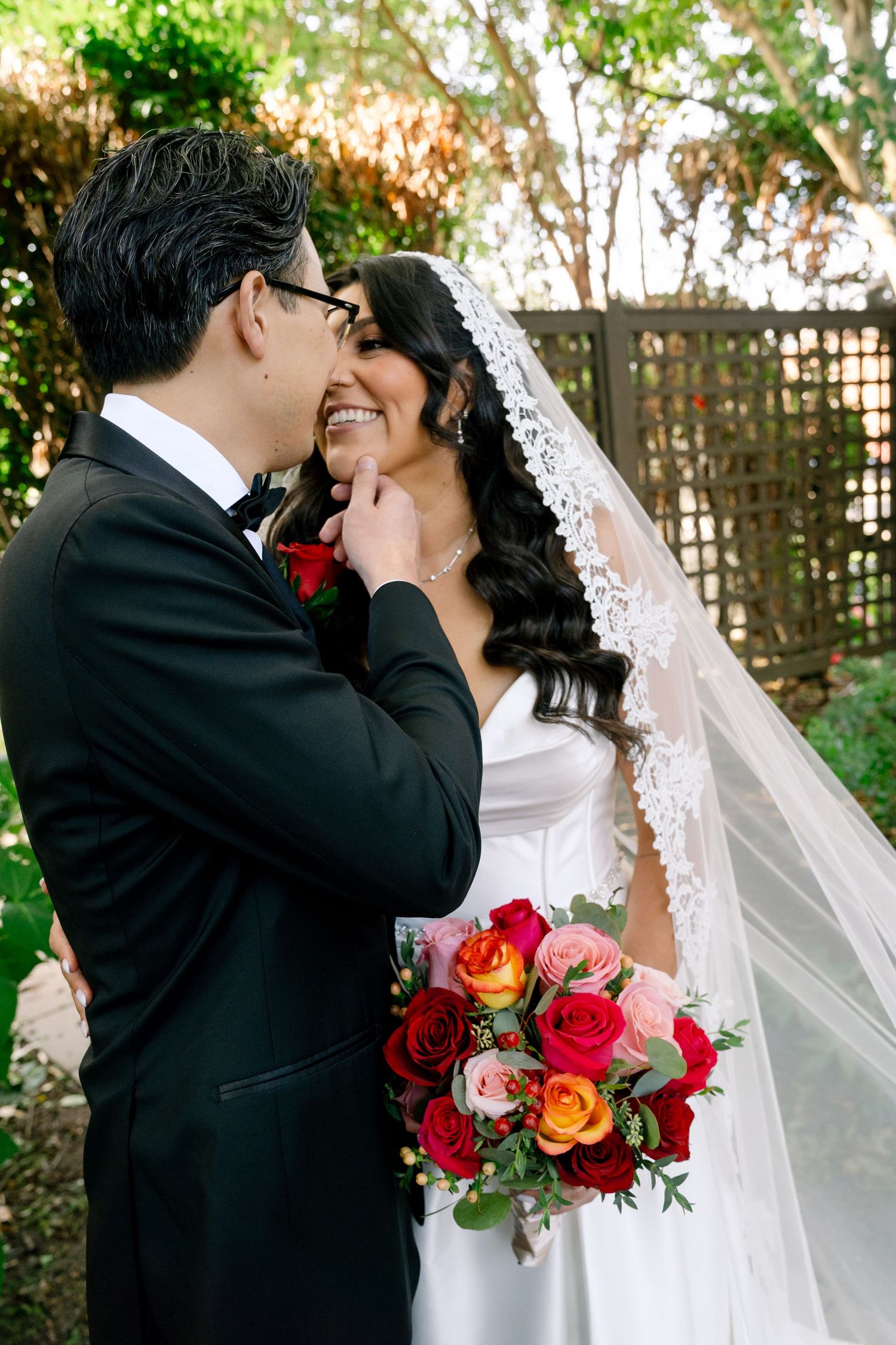 A romantic wedding moment in a garden setting with vibrant red and orange roses in the bouquet.