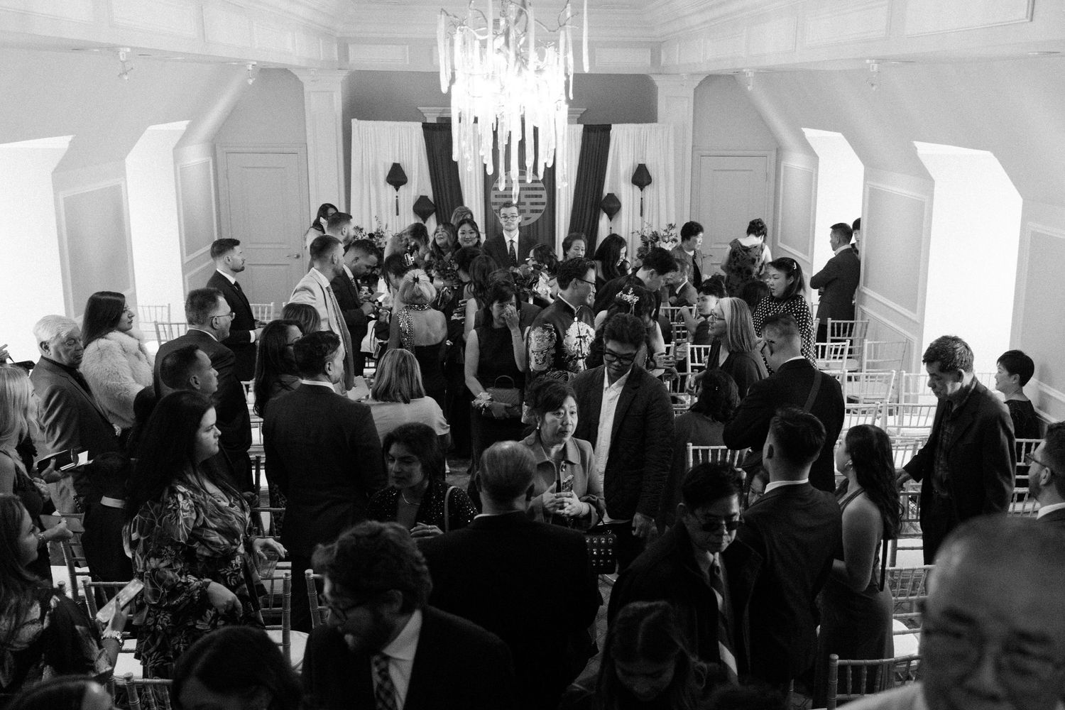 Black and white photo of wedding reception with guests seated under grand chandelier.