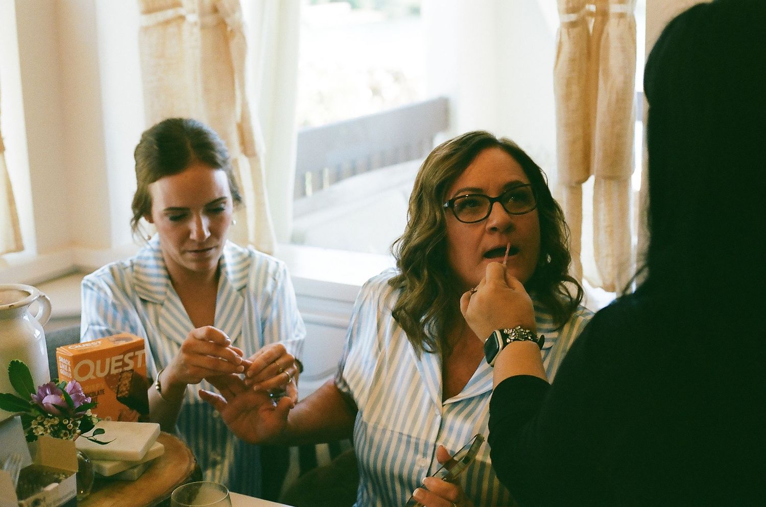 Two people sitting at a table enjoying snacks and drinks in a sunlit room with curtained windows.