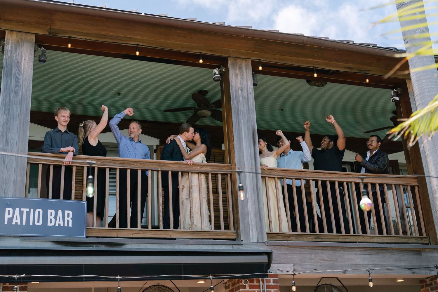 Group of people celebrating on a patio bar balcony with arms raised in cheer.