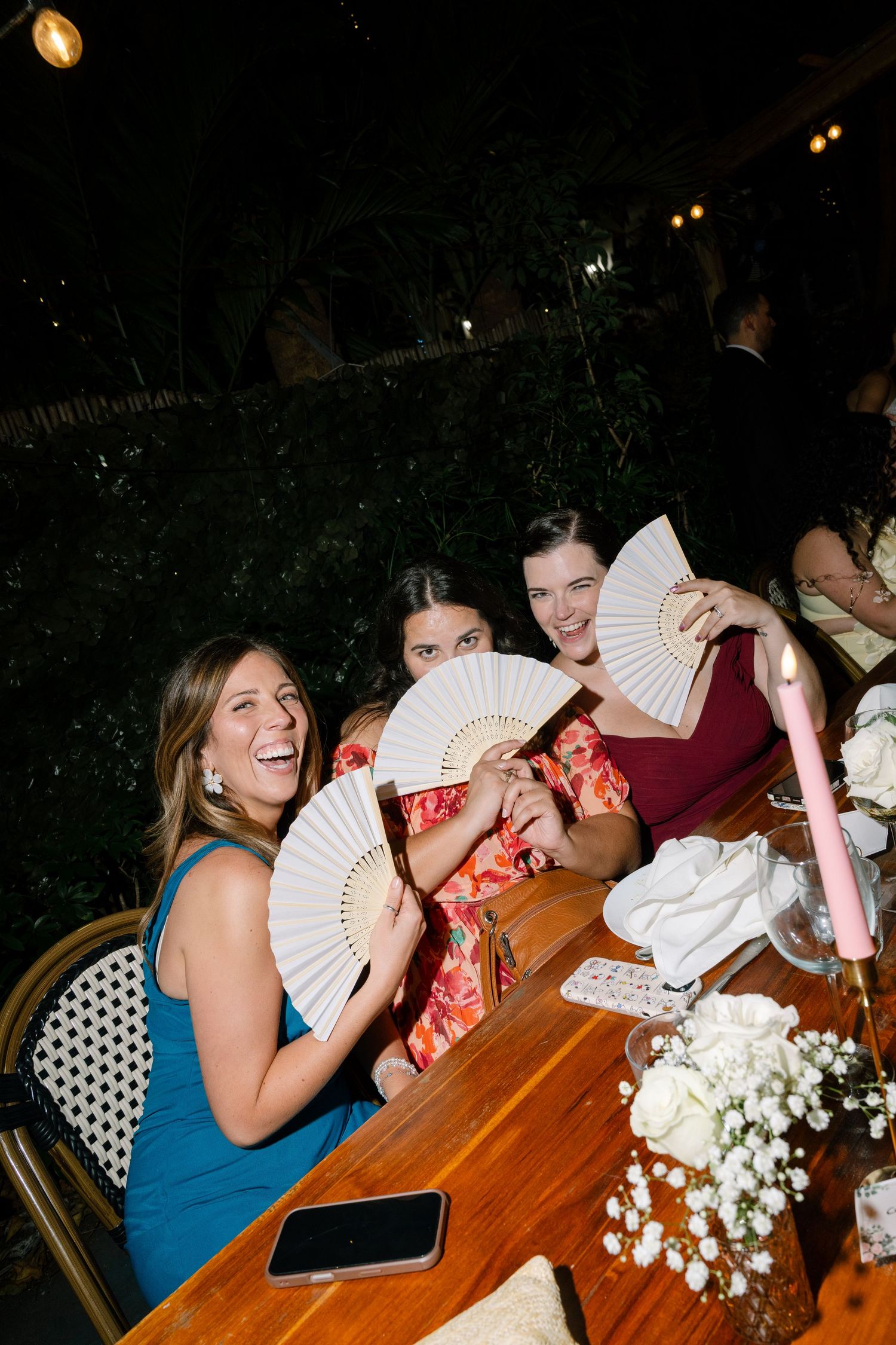 Women laughing and holding white hand fans at an outdoor wedding reception dinner table with white floral centerpieces.