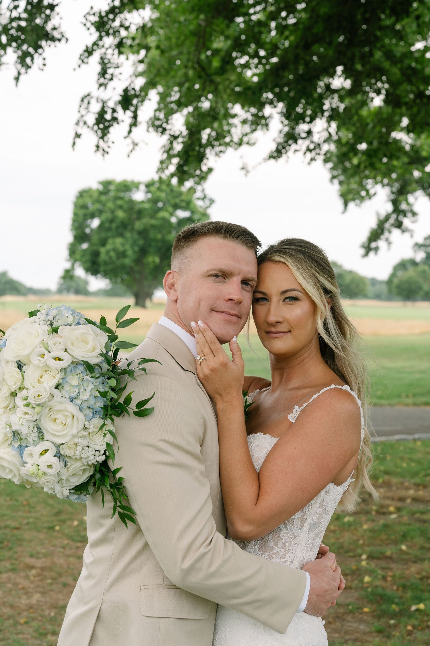 A bride and groom embrace outdoors under a tree, she holds white and blue roses, both smiling in wedding attire.