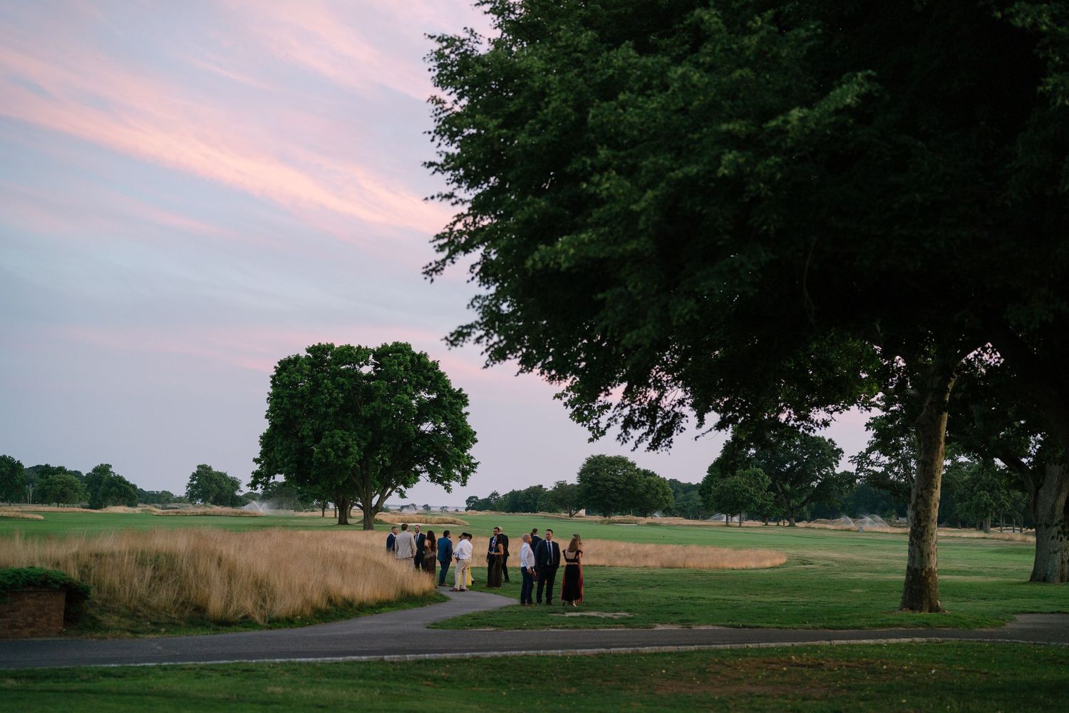 Group of people gathered on a golf course path at dusk with trees and pink sky in background.