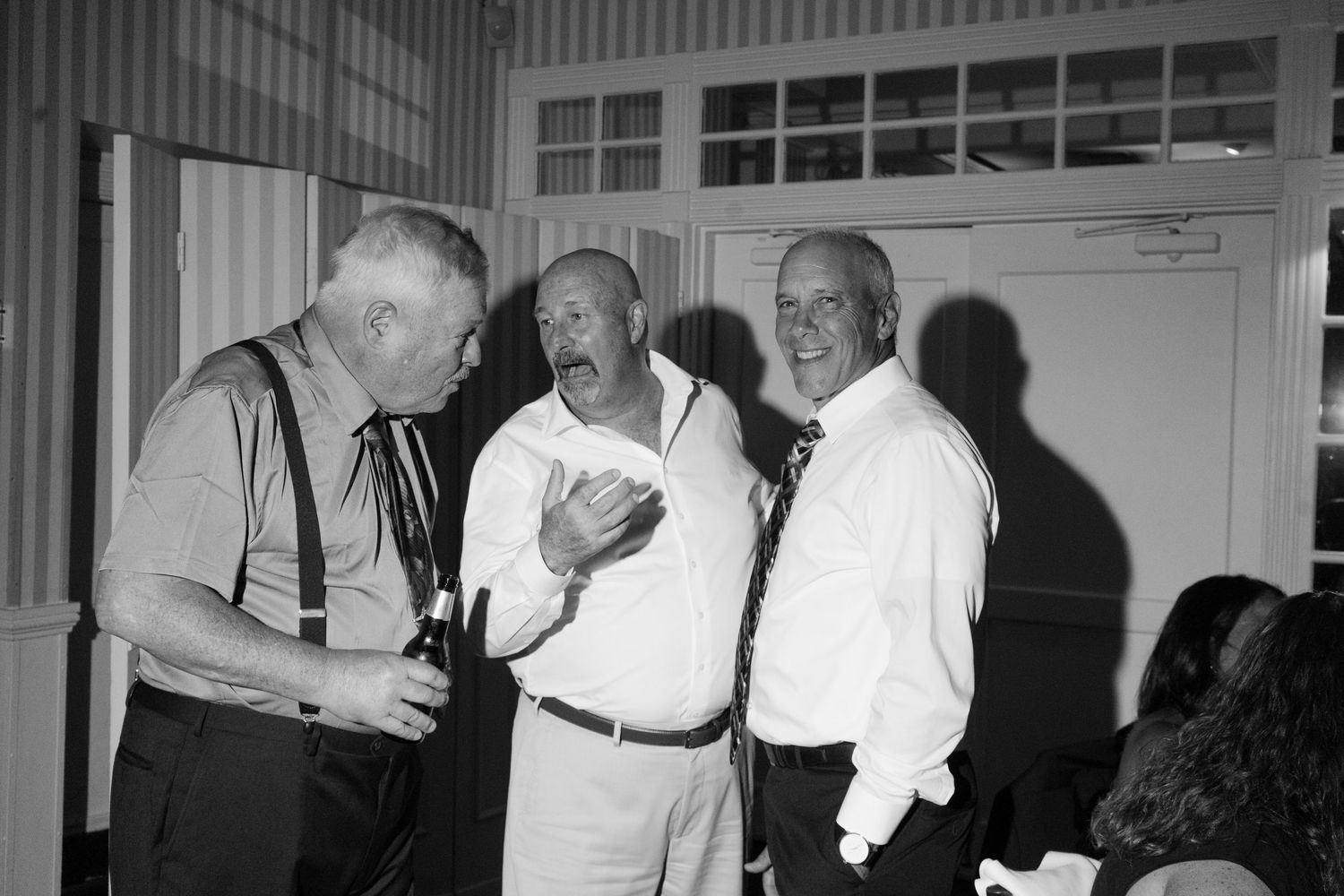 Three older men in formal attire converse together in a black and white candid photo.