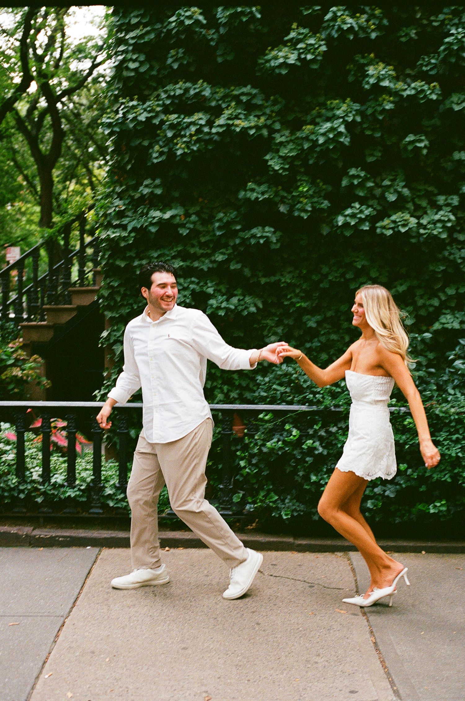 A joyful couple in white outfits dancing and holding hands on a sidewalk surrounded by lush green ivy walls.