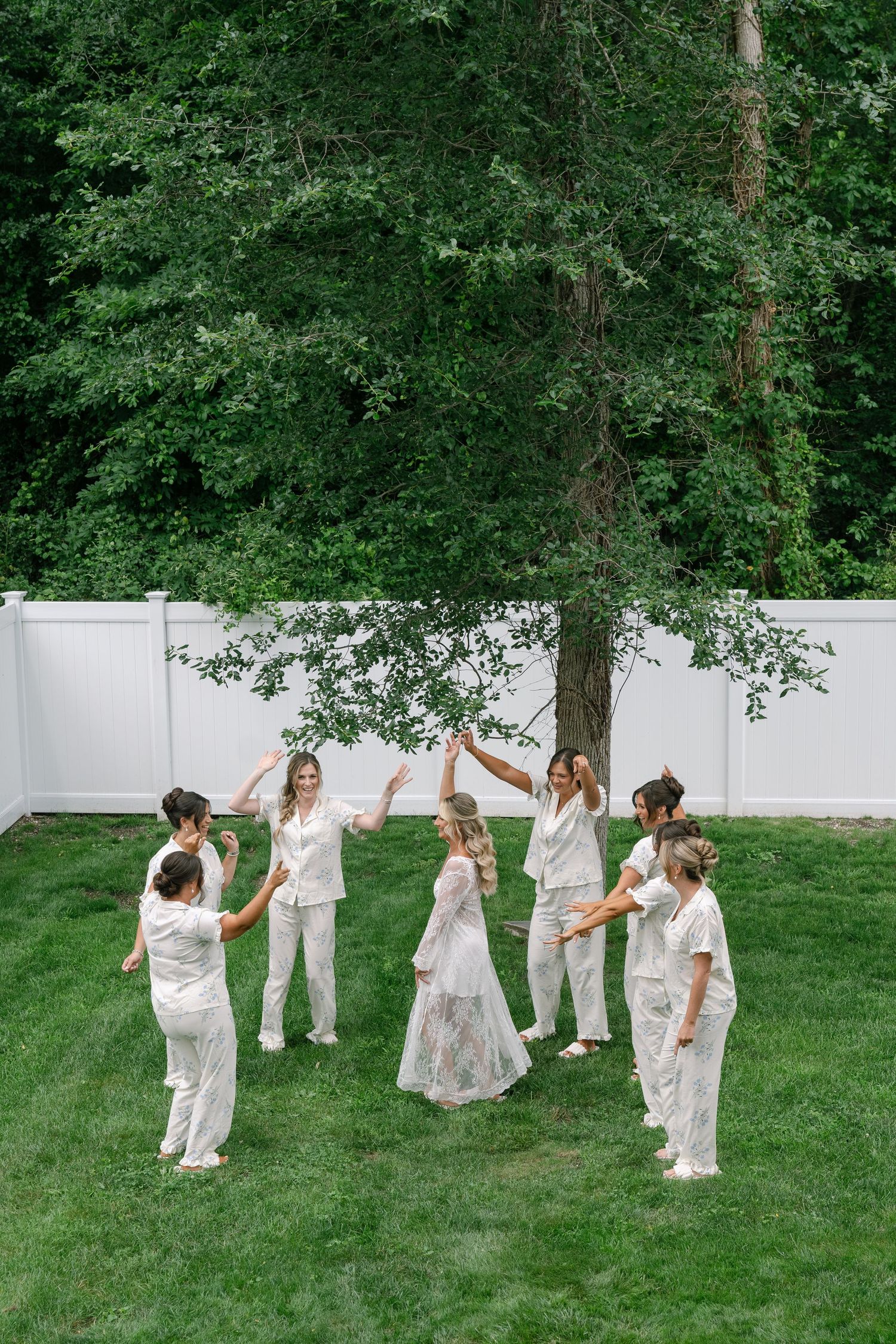 Bride in lace gown surrounded by bridesmaids in white pajamas celebrating outdoors on green lawn with trees and white fence.