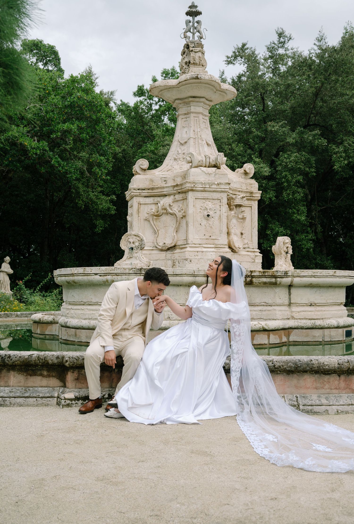 A couple shares a romantic moment by an ornate stone fountain surrounded by green trees in an elegant garden setting.
