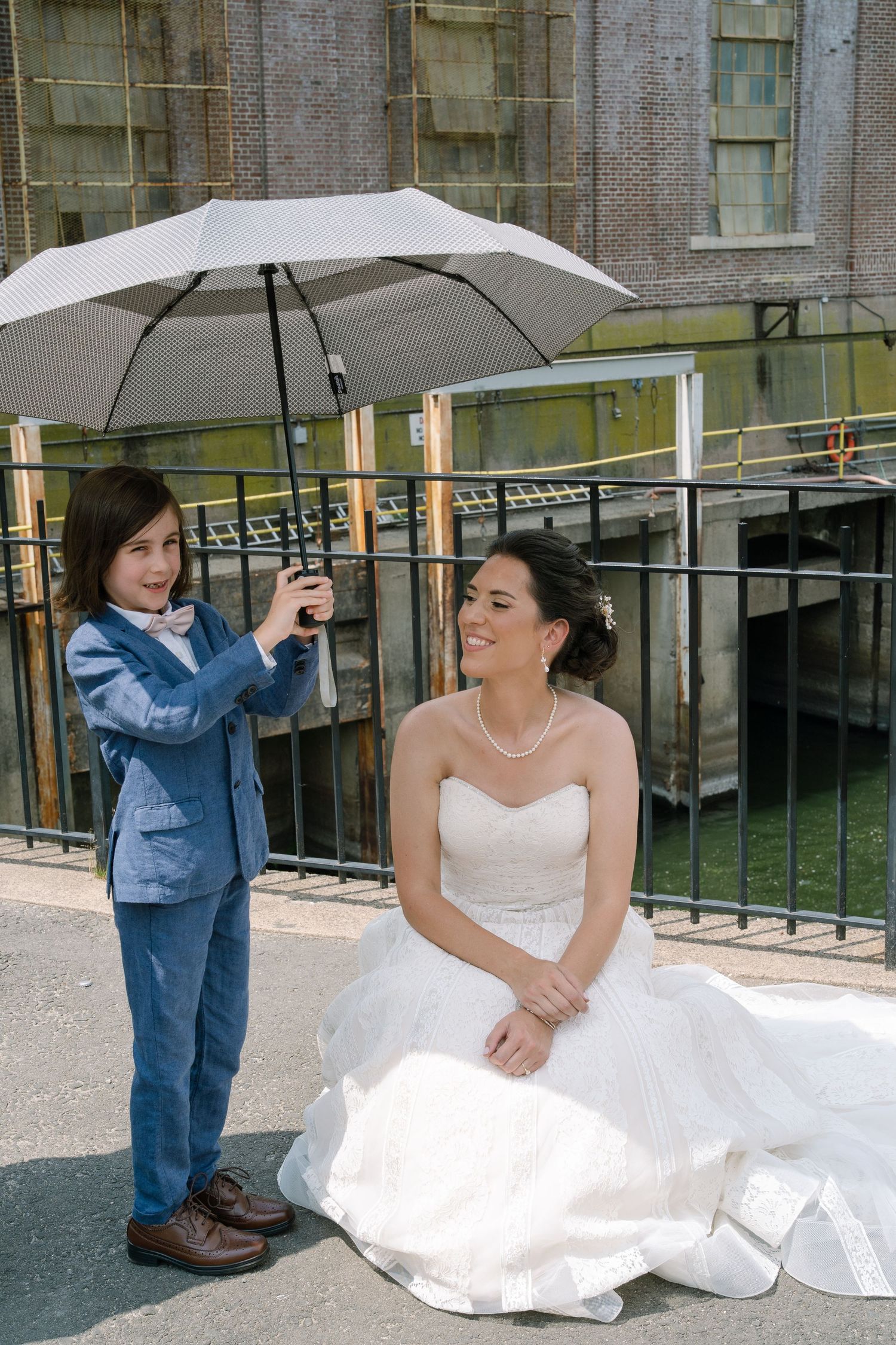 Someone holds a white umbrella over a person in a white wedding dress sitting outdoors by a metal railing.