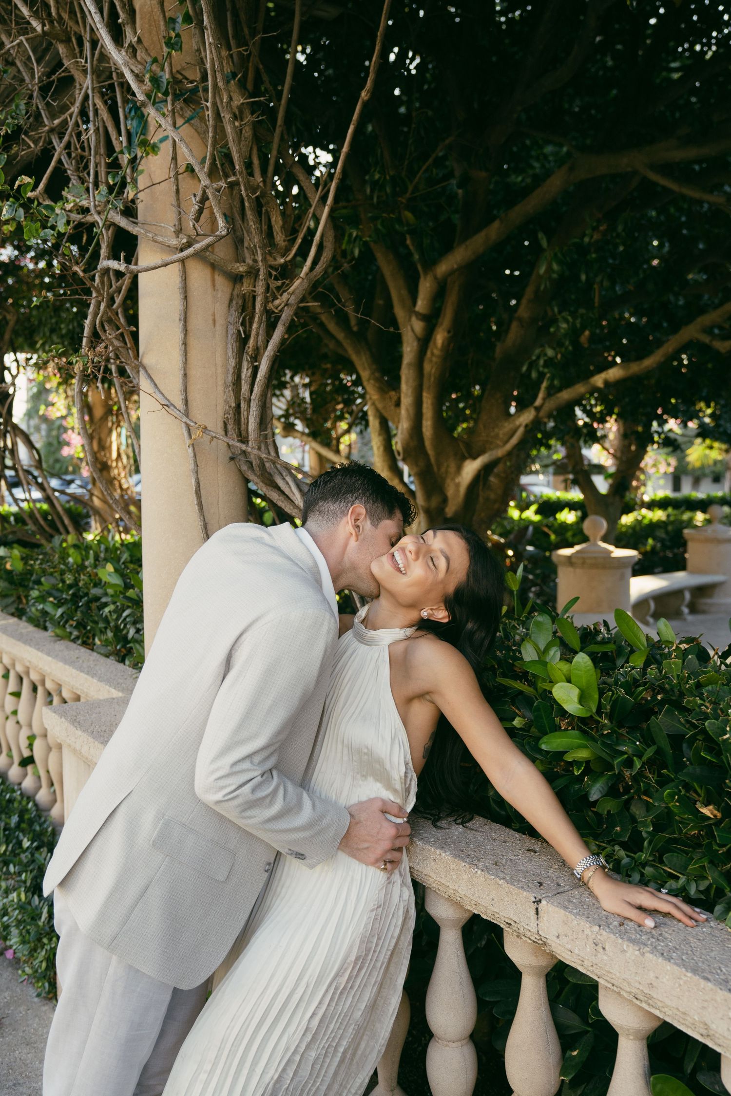 Two figures share an intimate moment leaning against a stone railing surrounded by lush greenery and trees in a garden setting.