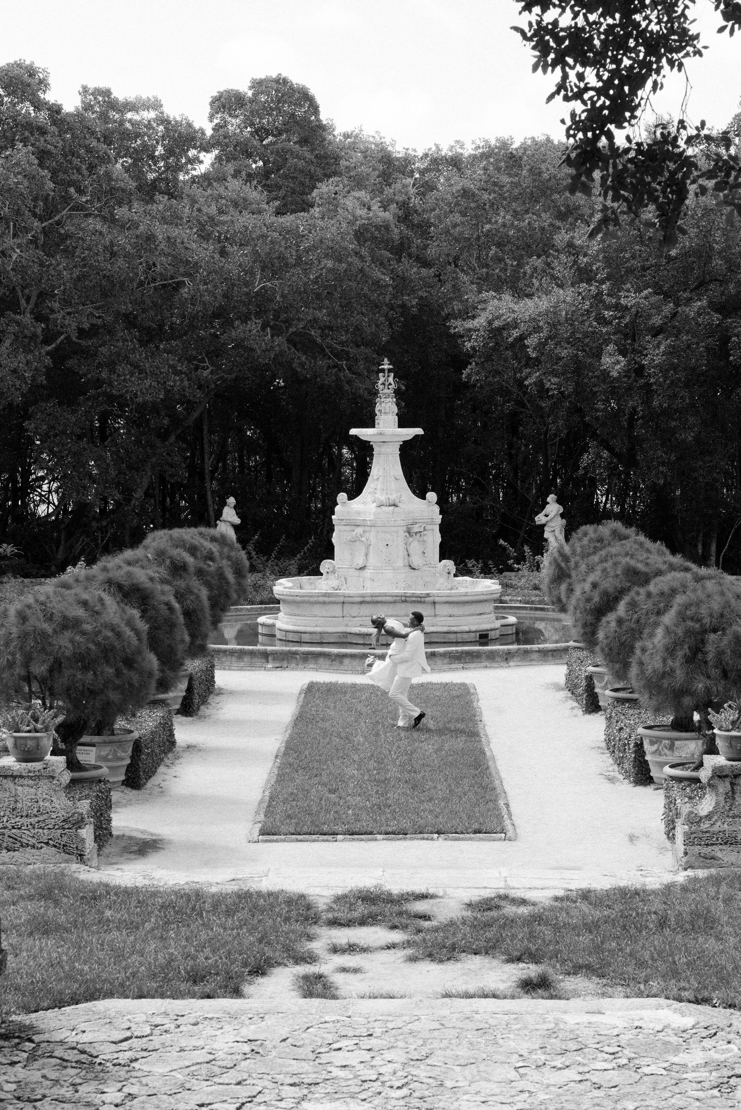 A series of ornate white marble fountains and decorative elements arranged along a formal garden path lined with hedges.