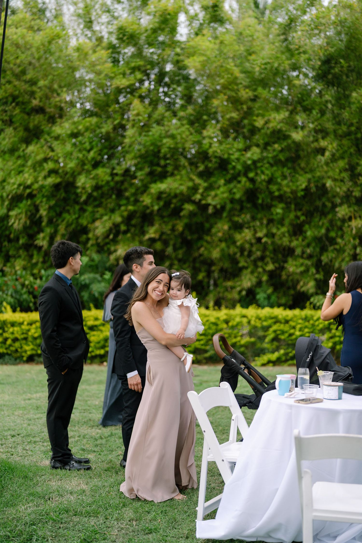 A bridesmaid in a taupe dress holds a young child at an outdoor garden wedding reception.