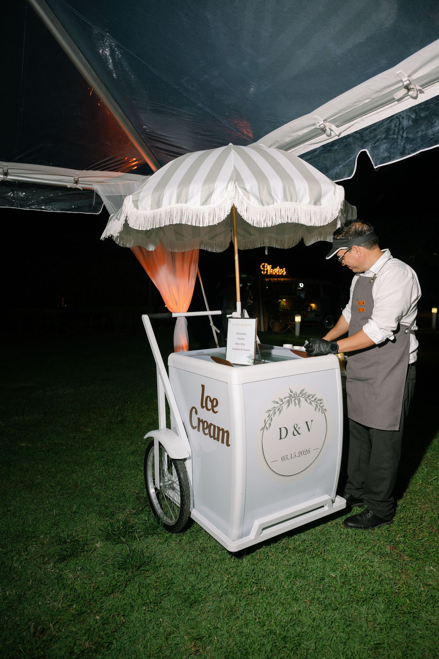 Elegant white ice cream cart with striped umbrella and D&V monogram at an outdoor wedding reception at night.