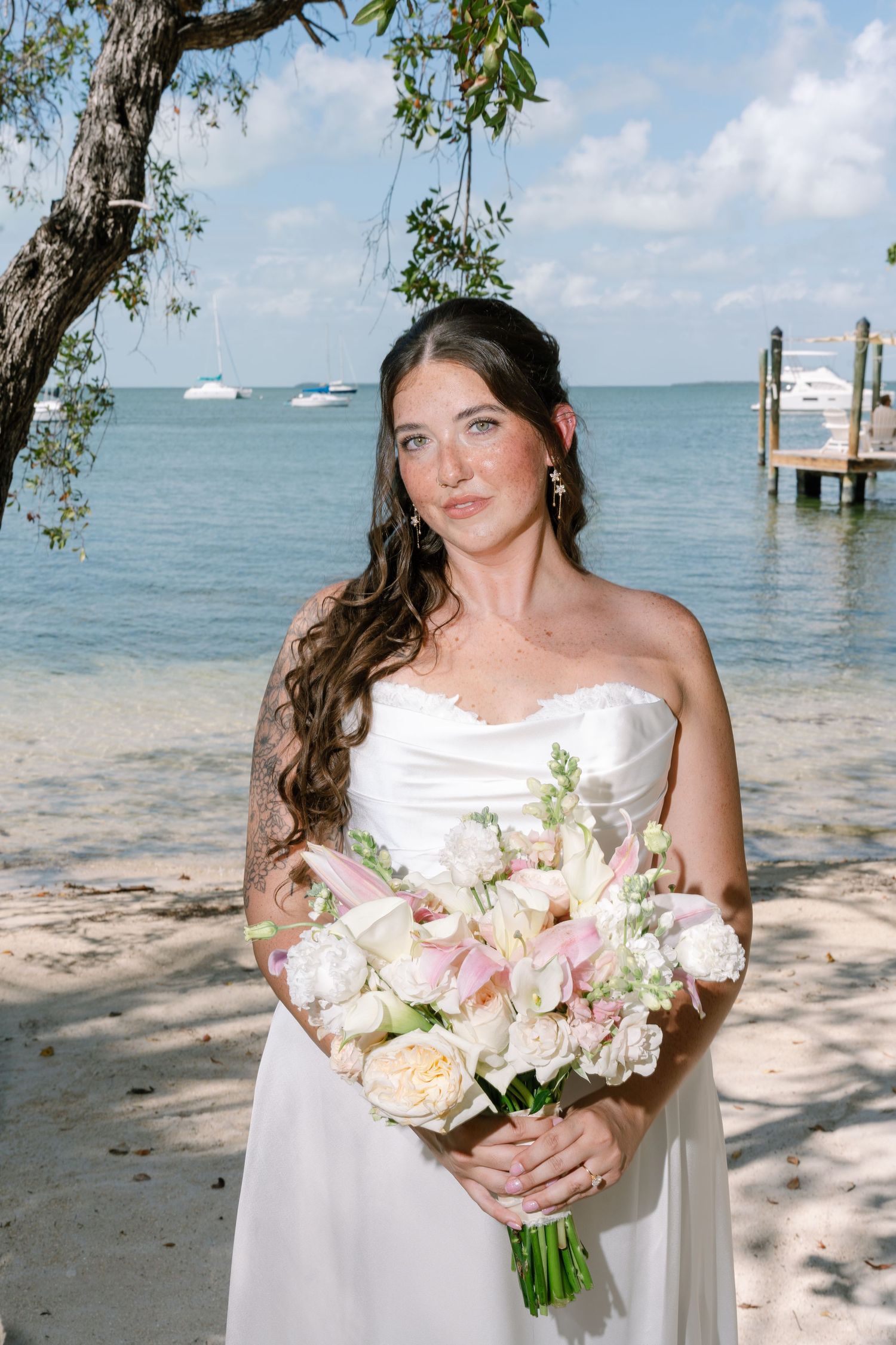 A woman in a white wedding dress holding a bouquet of flowers on a beach with boats in the background.
