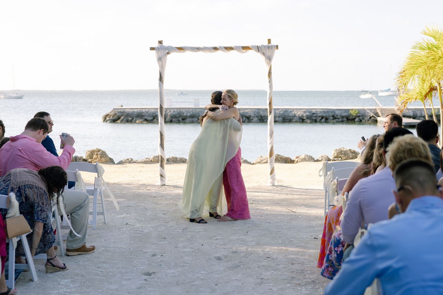 Two brides embrace under a wooden arch at a beachside wedding ceremony with guests seated on white chairs.