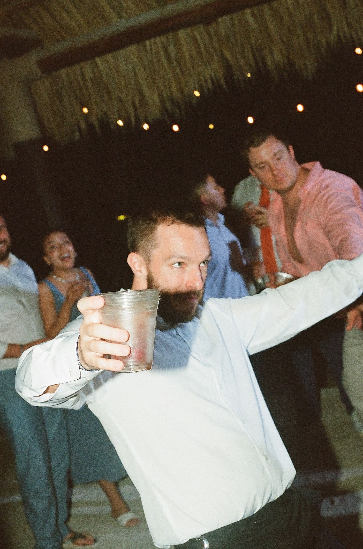 Bearded wedding guest raises cup in toast while dancing at outdoor tropical reception.