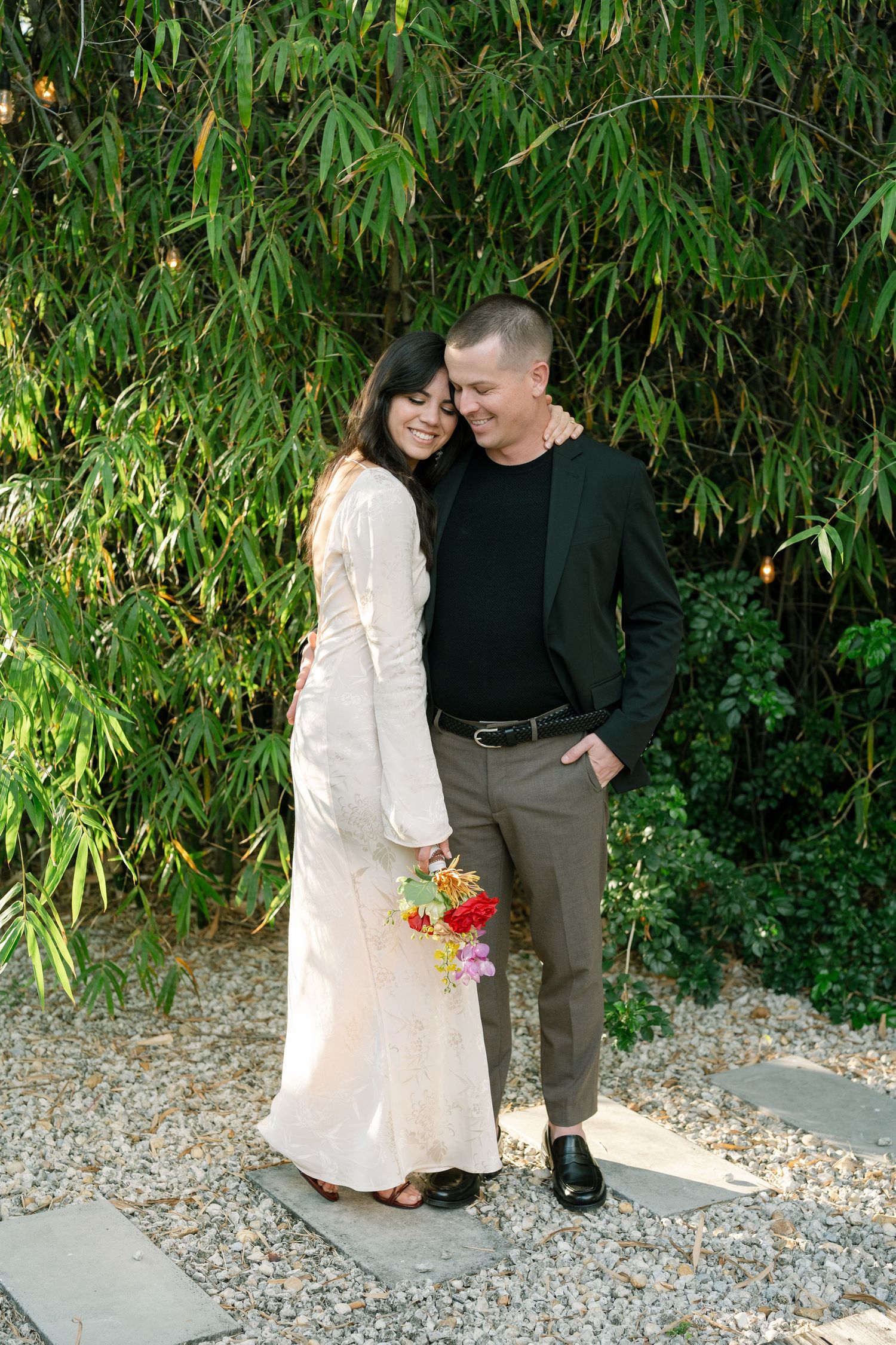 A couple embraces on a gravel pathway surrounded by bamboo and tropical greenery.