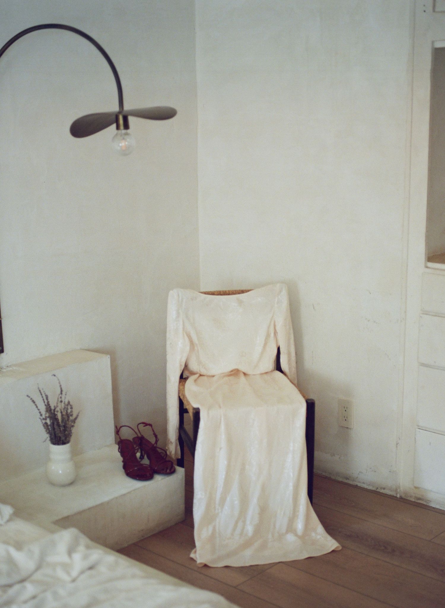 A minimalist room corner with a white draped chair, arched floor lamp, and purple vase with dried flowers.
