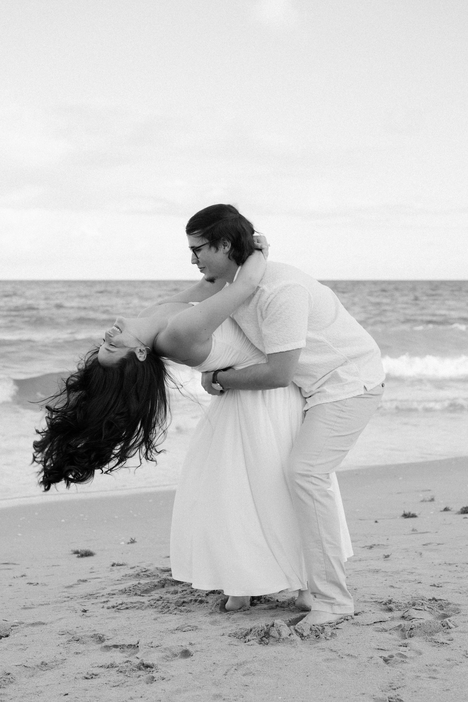 A couple in white clothing dances together on a beach in a romantic sequence of black and white photos.