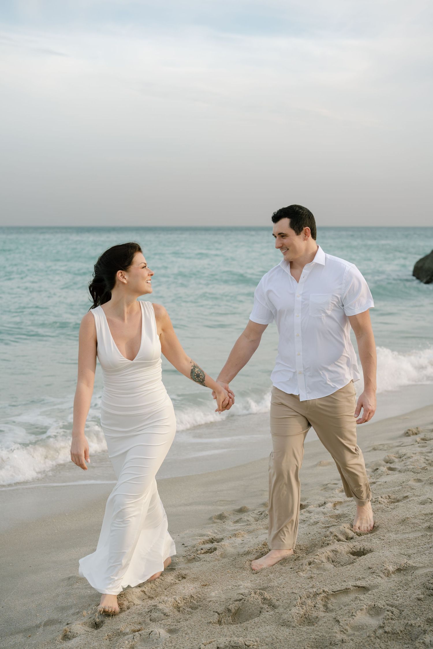 A couple walks hand in hand along a scenic beach shore at dusk wearing white and beige casual attire.