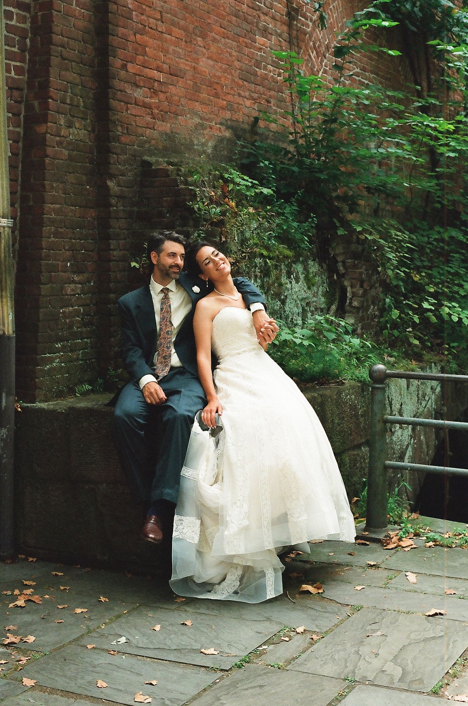 A bride and groom sit together on a brick wall surrounded by greenery during a wedding photoshoot.