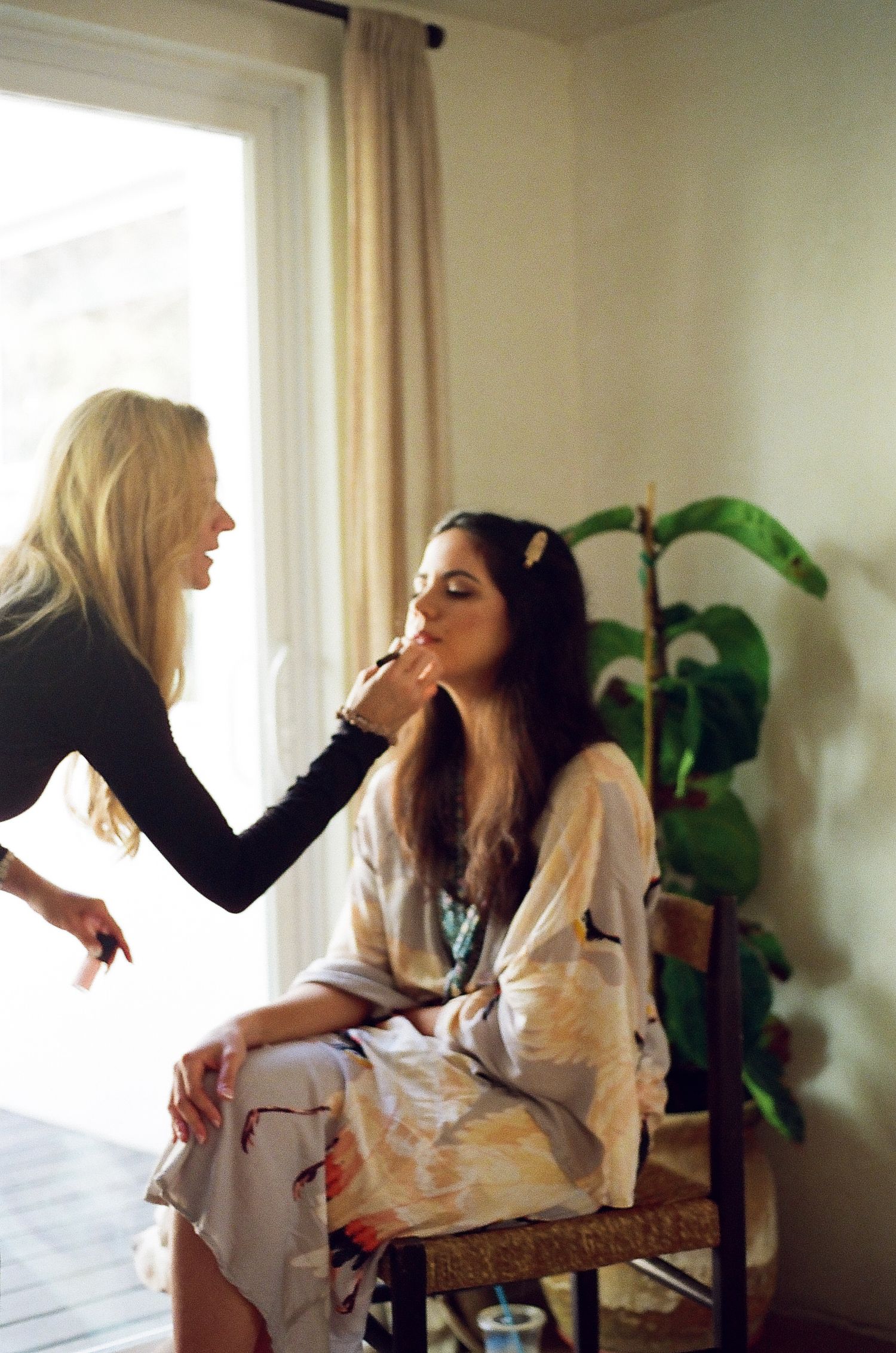 Two people in white clothing share a candid moment by a window with natural light streaming in.