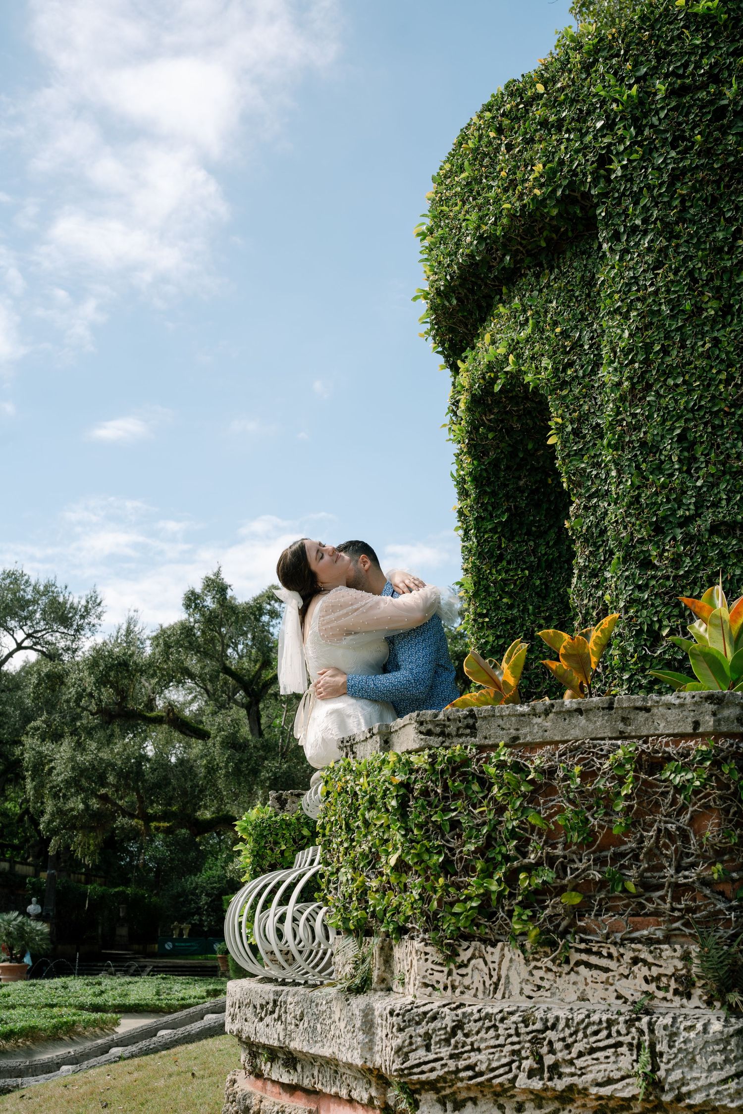 A romantic moment captured on a stone wall with ivy-covered backdrop and yellow lilies in the garden.