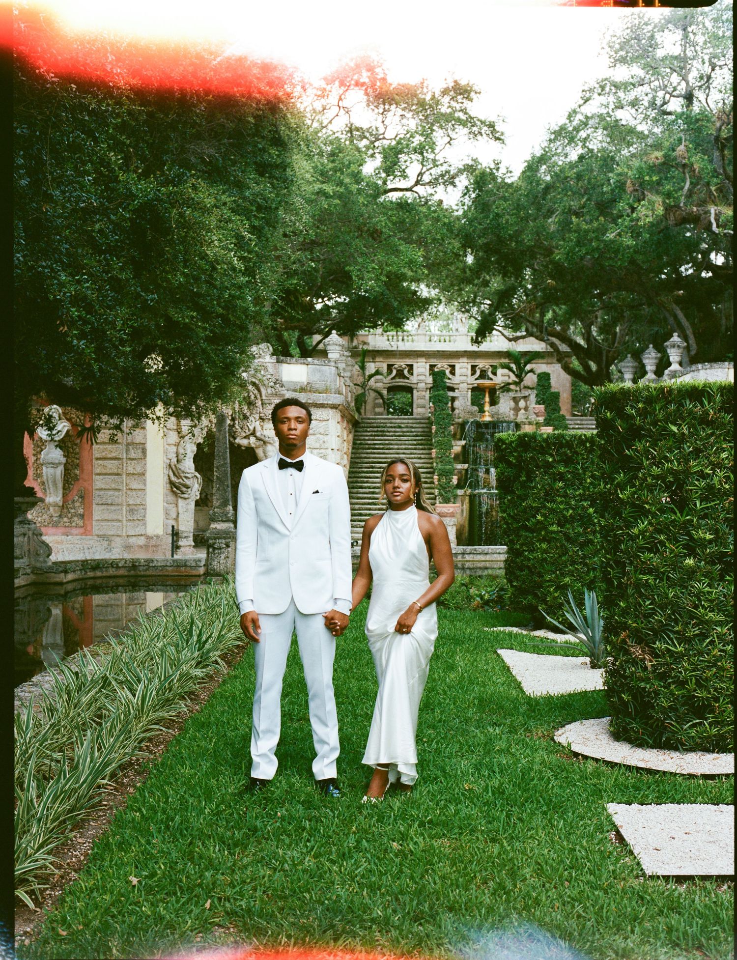 A couple in elegant white attire poses in a lush garden setting with ornate stone architecture and stepping stones.