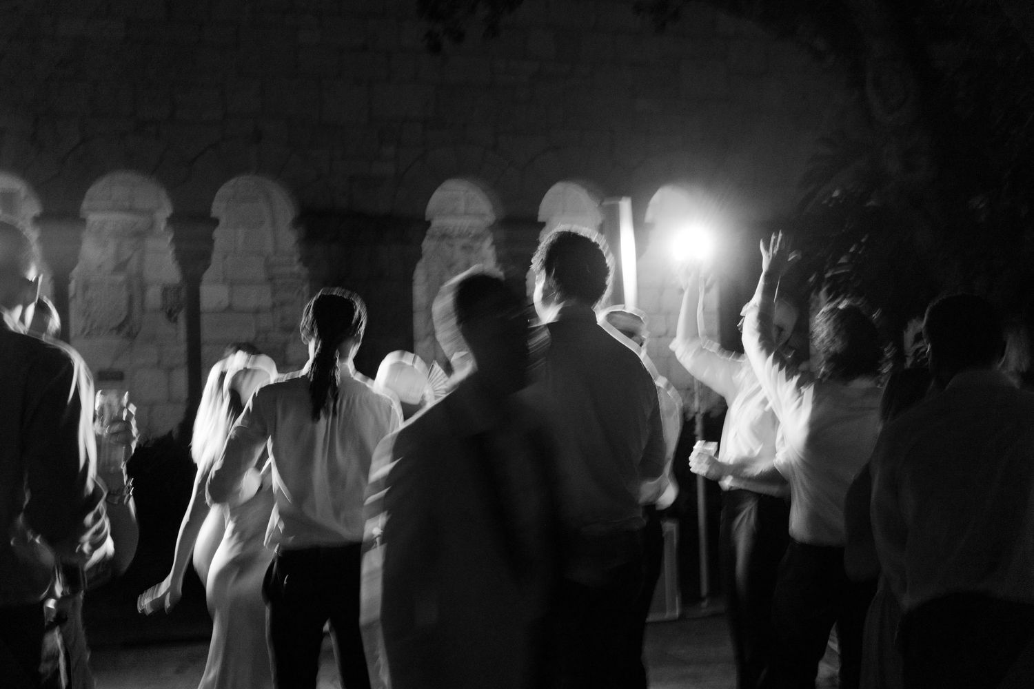 Black and white photo of wedding guests dancing at night in stone arched venue.