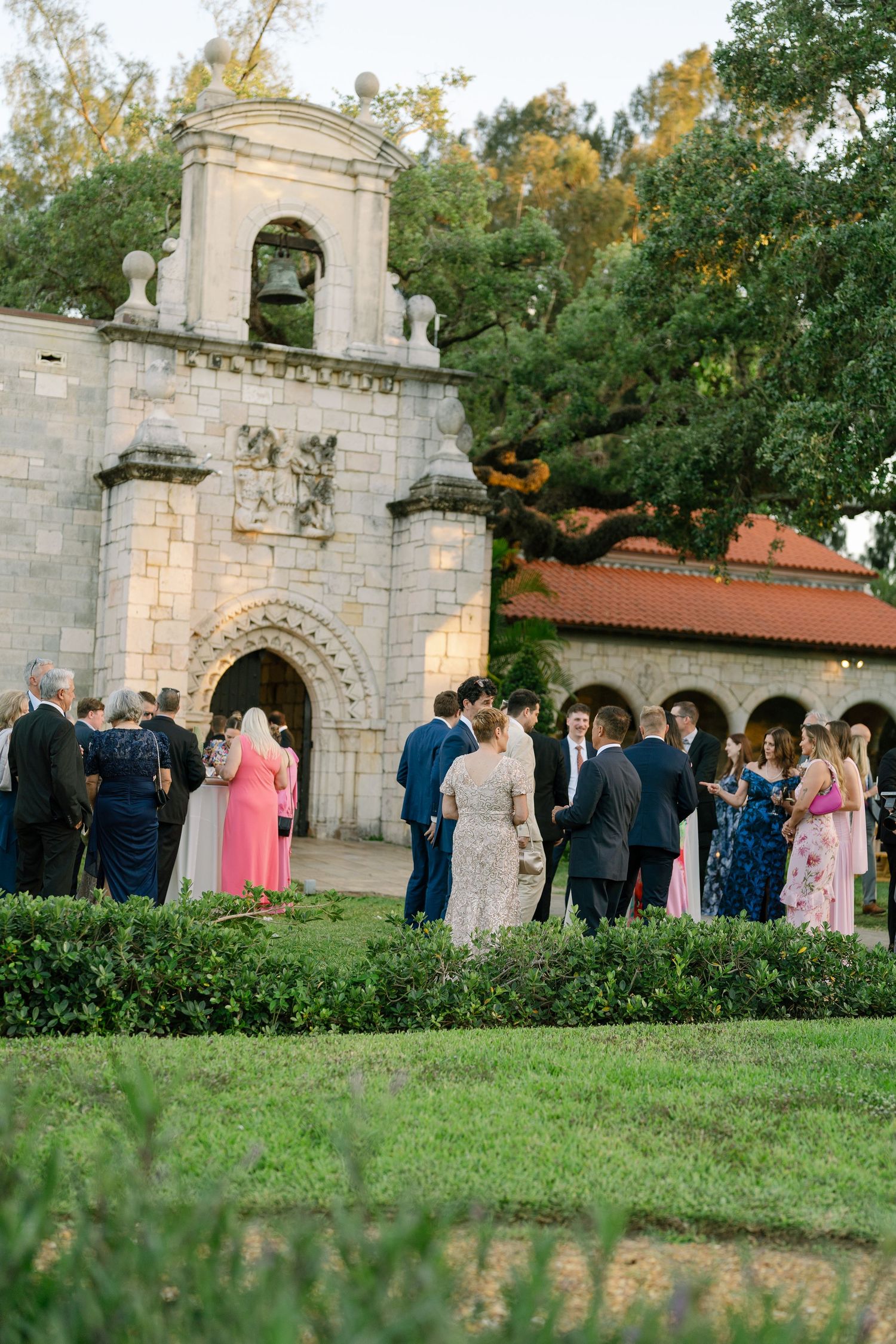 Wedding guests mingle outside a historic stone chapel with bell tower surrounded by lush greenery at golden hour.
