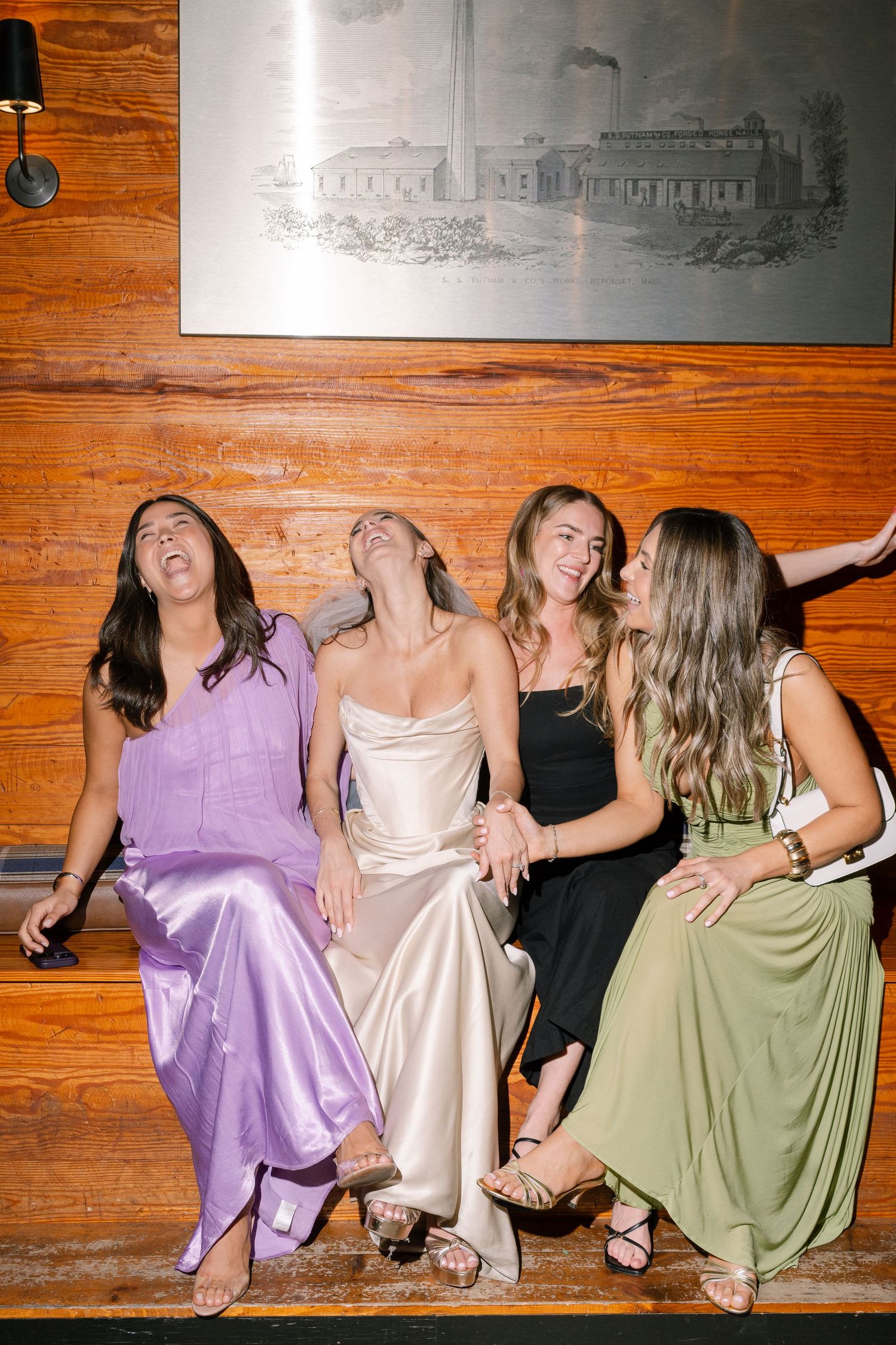 Four women in formal dresses laughing together on a bench at a wedding reception.