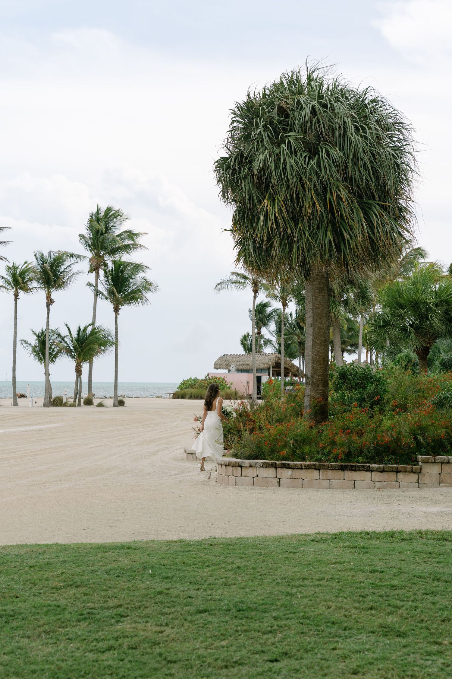 Woman in white dress walks near tropical palm trees on sandy beach resort path.