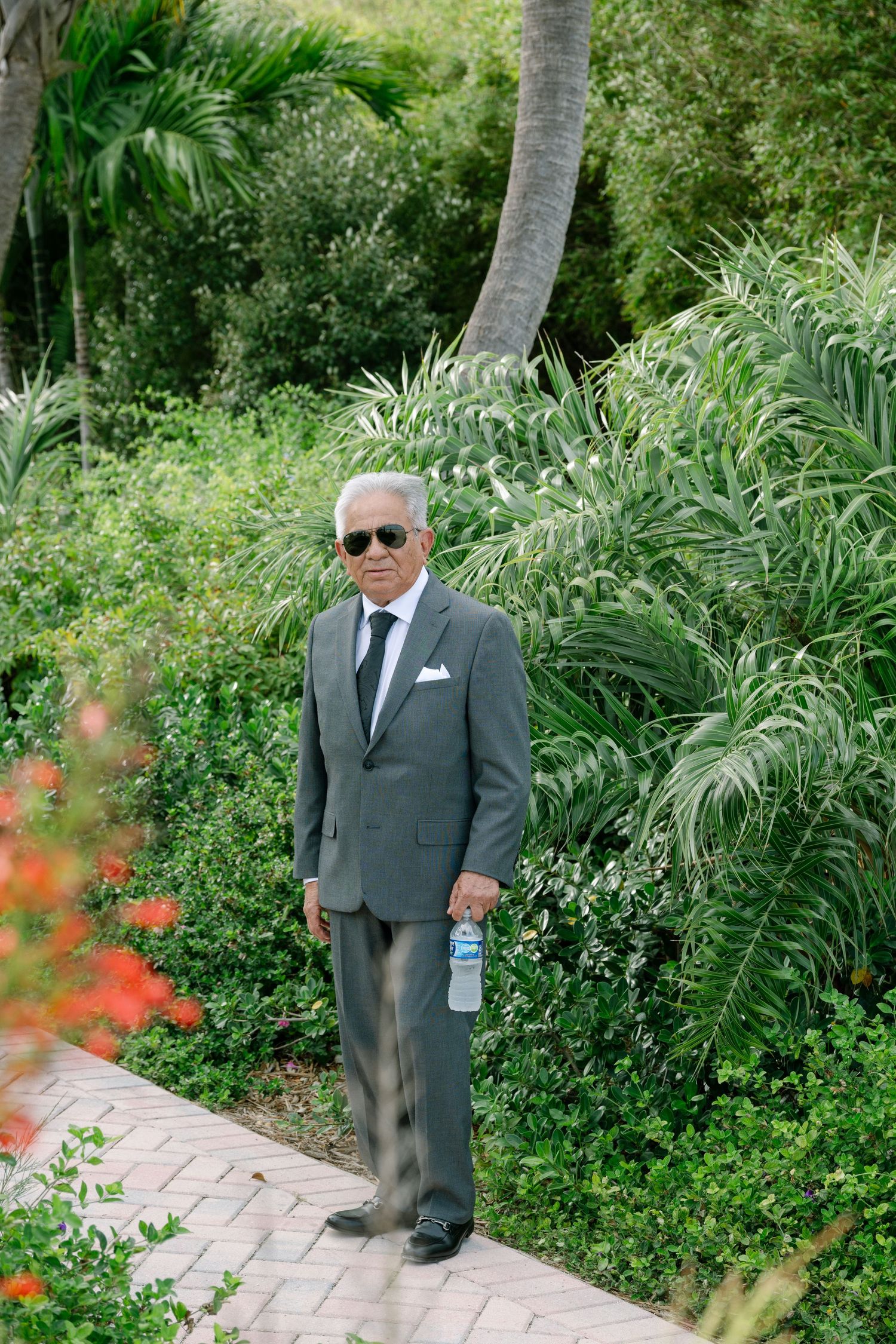 Elegant older gentleman in gray suit with sunglasses holding water bottle on brick garden path.