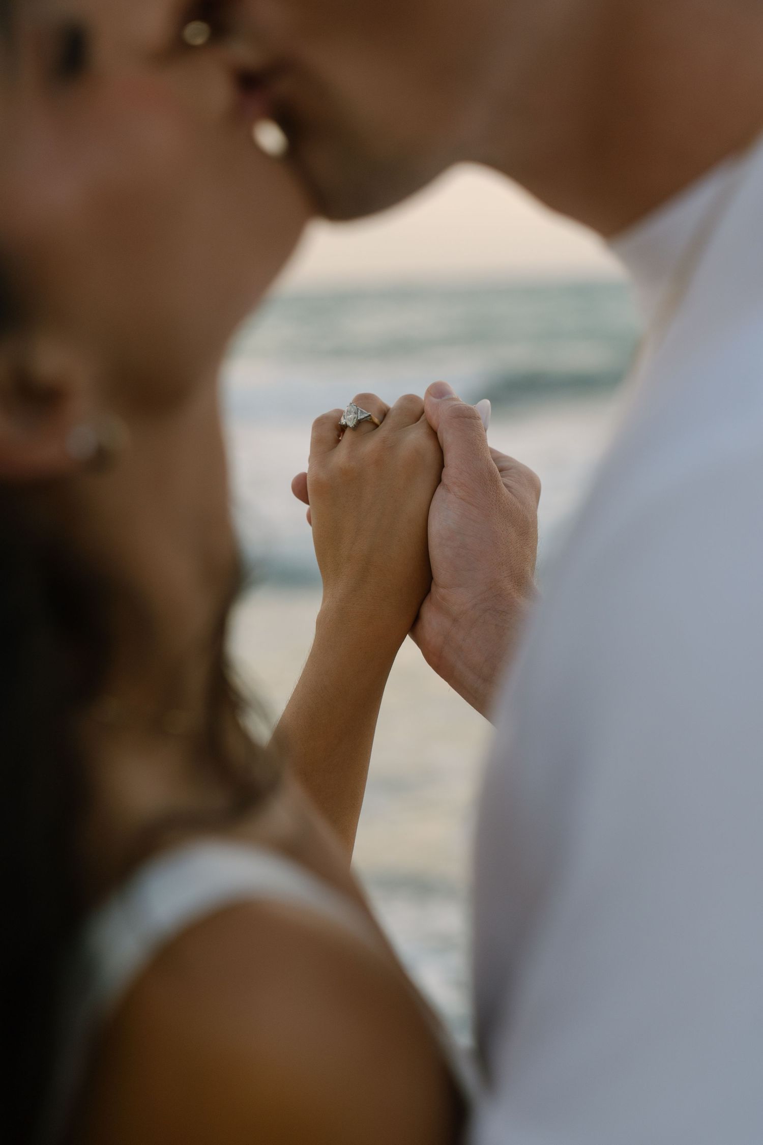 Couple kissing on beach at sunset, holding hands with engagement ring visible in foreground.