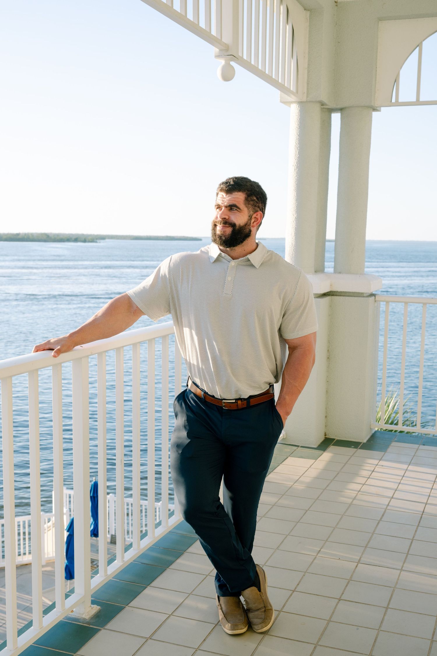 A man in casual attire leans against a white railing on a waterfront porch overlooking the ocean.