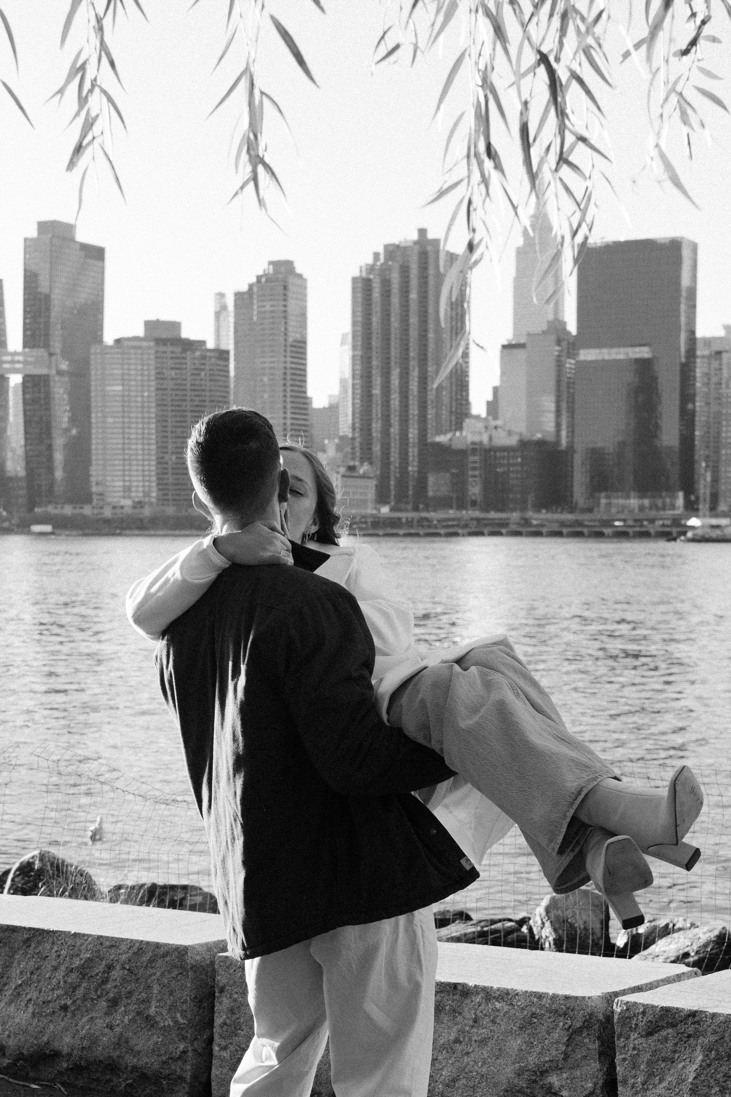 Black and white photo of two people gazing at the New York City skyline from a waterfront railing.