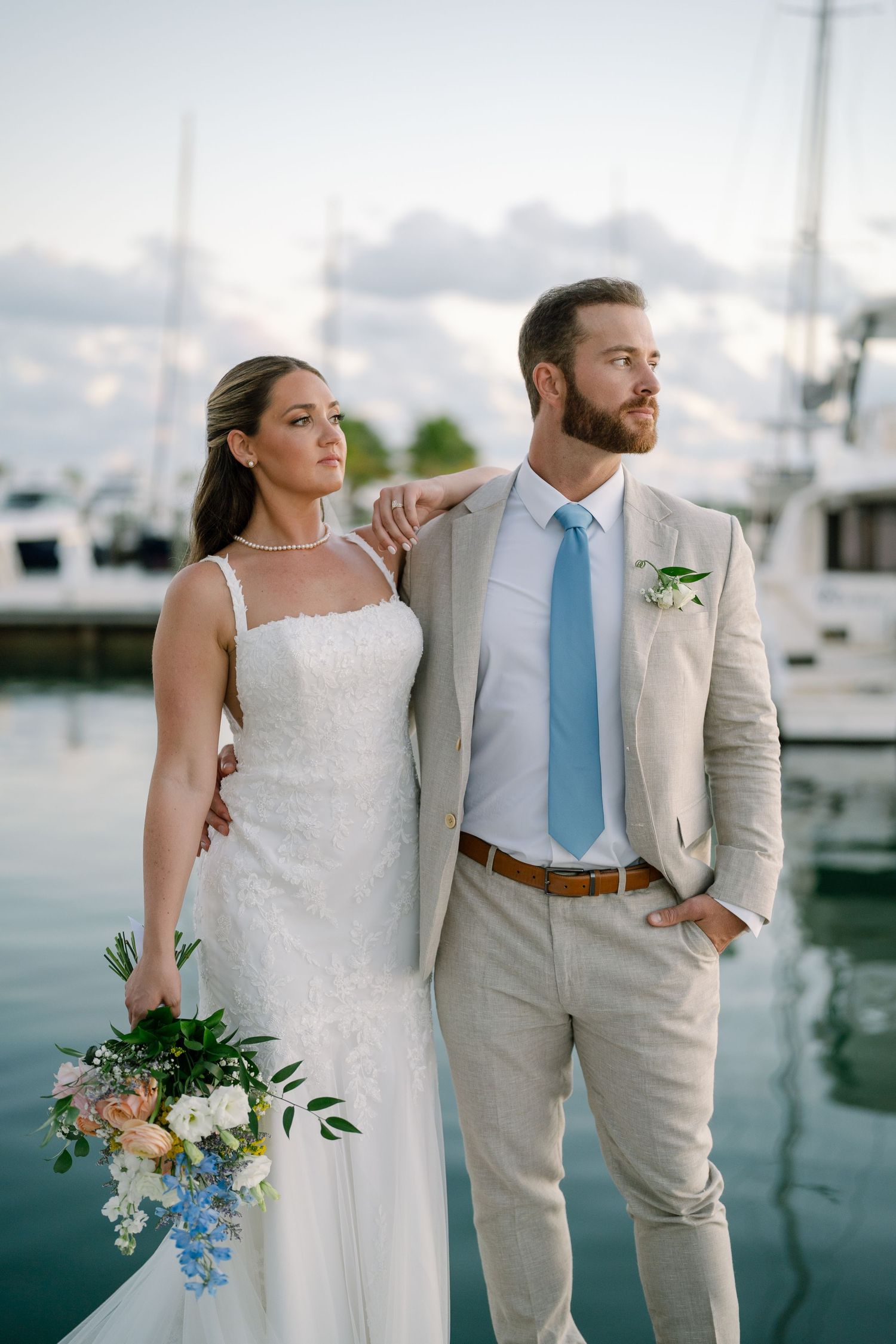 Elegant couple in wedding attire poses by marina with boats in background.