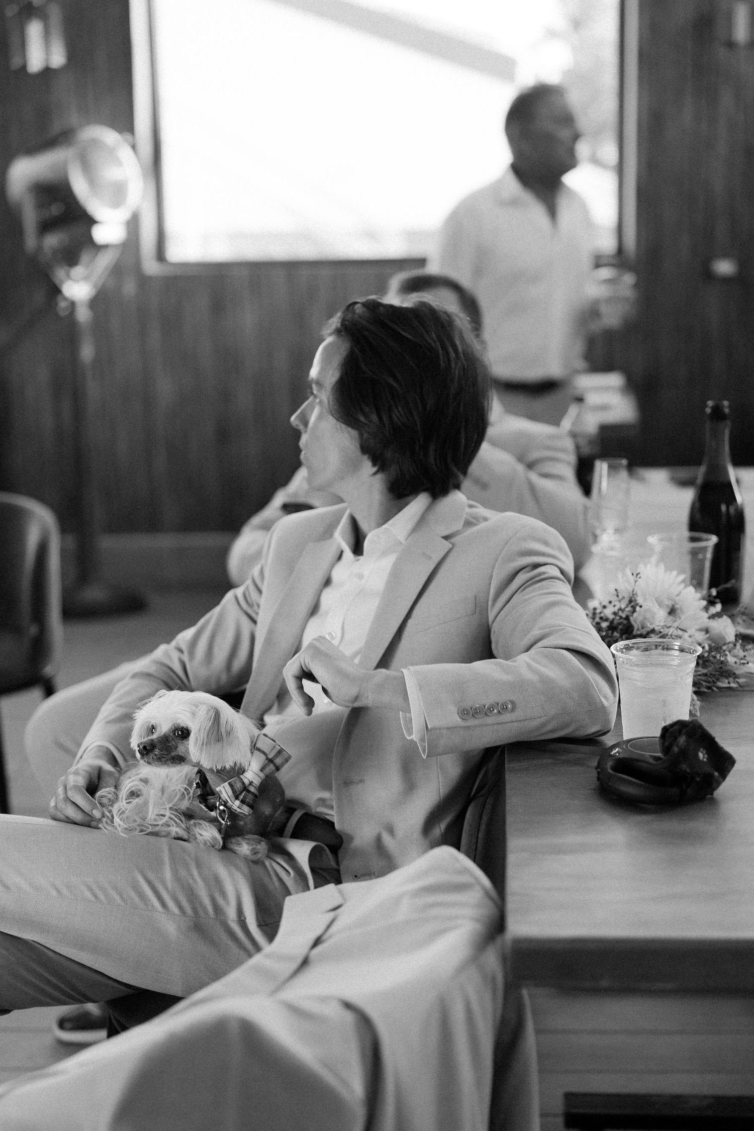 A black and white candid image shows someone in a light blazer relaxing at a table with drinks and bottles.
