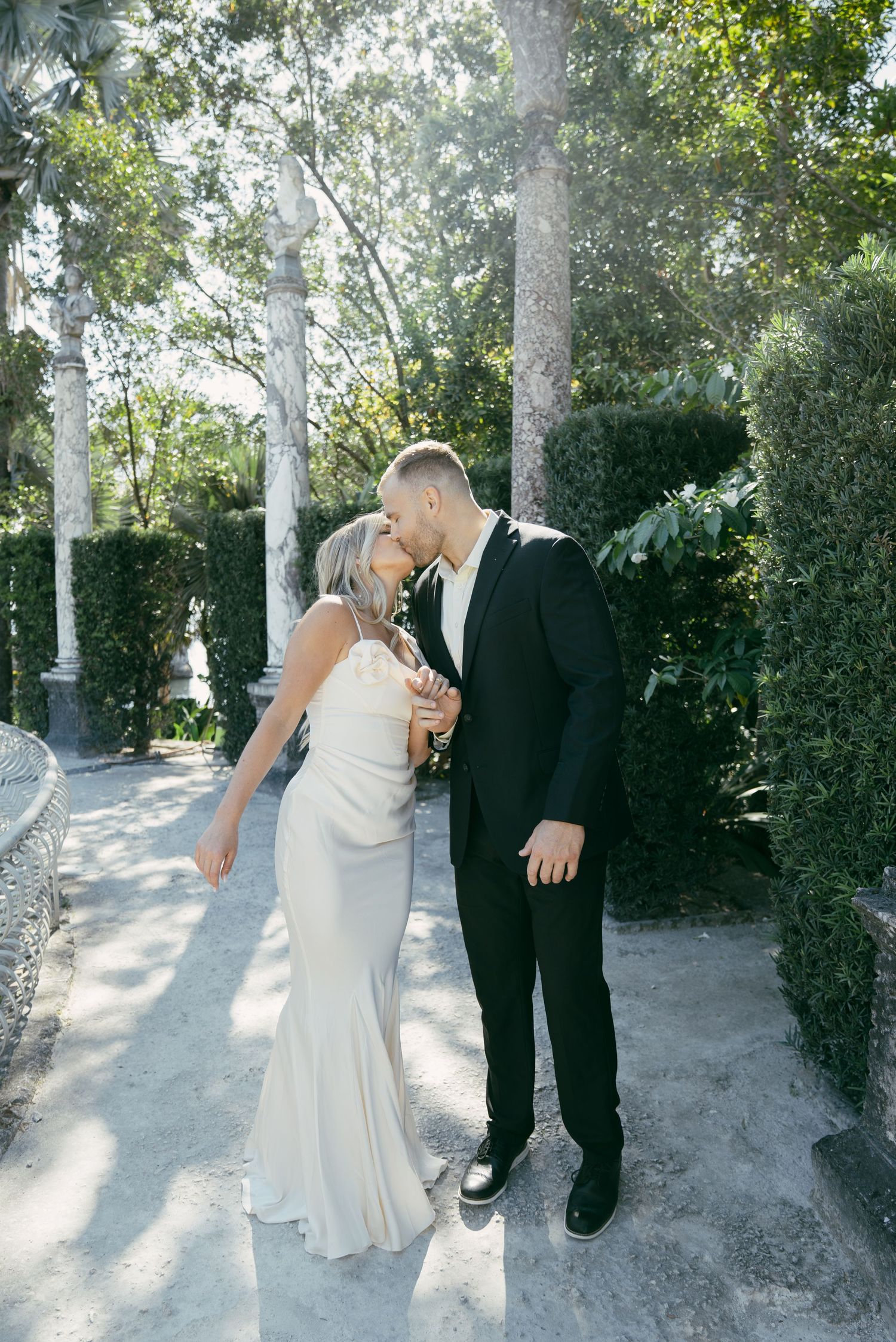 Elegant wedding couple walking together on a tree-lined path with soft sunlight filtering through the branches.