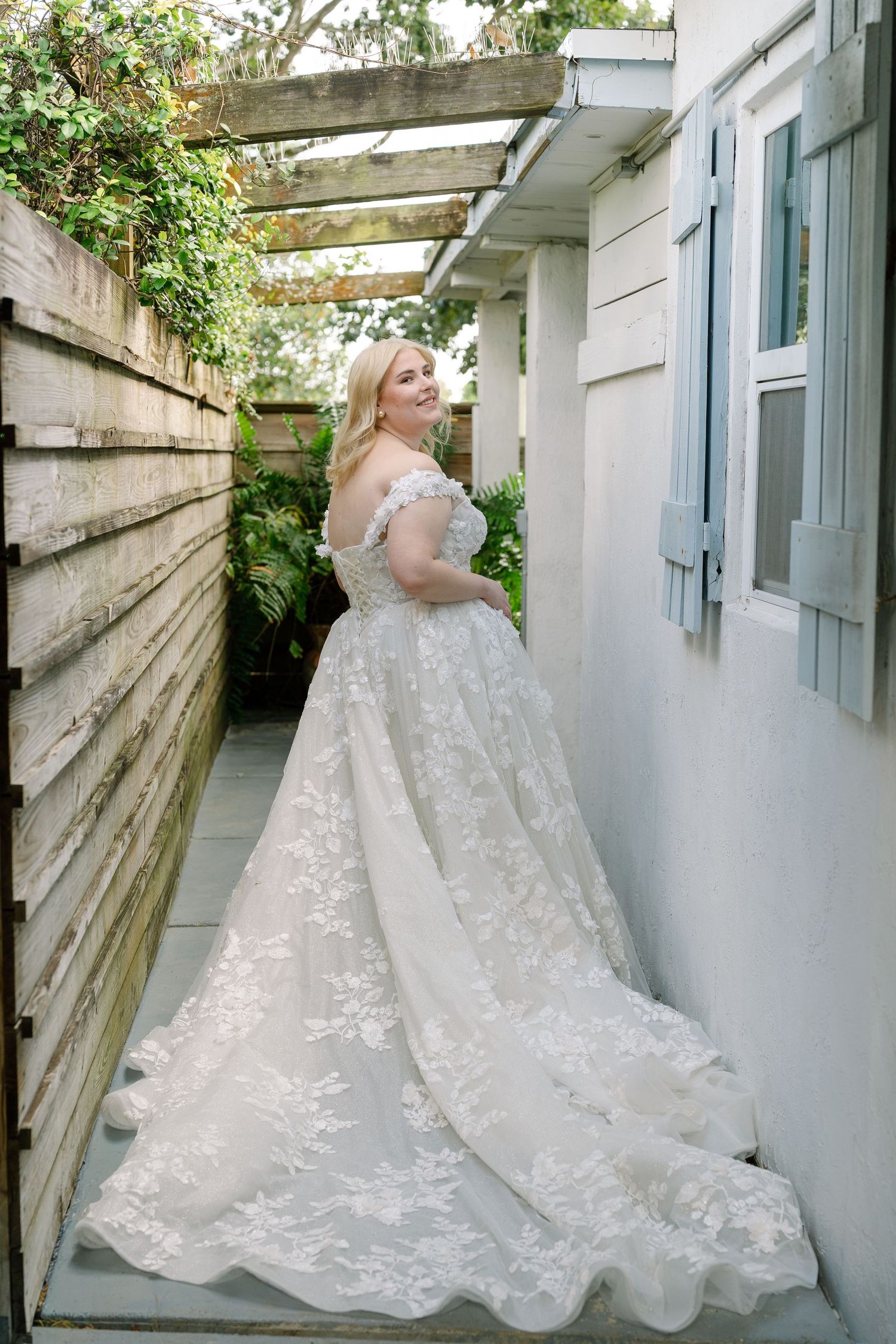 A woman wearing a beautiful white lace wedding dress stands in a garden with a wooden fence and greenery in the background.