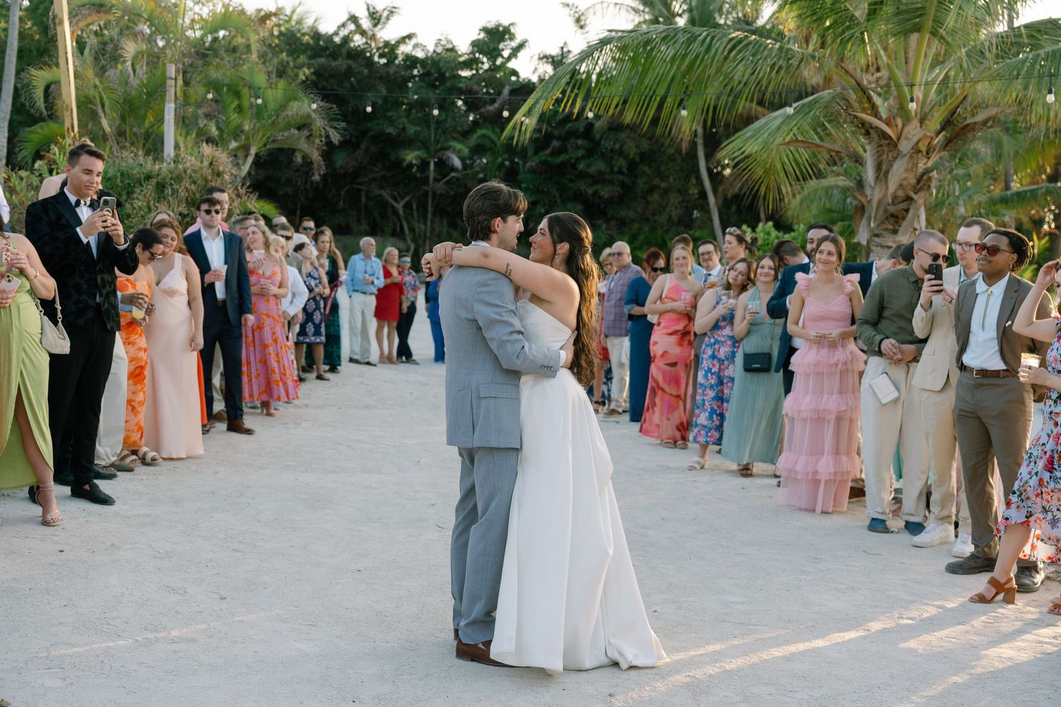 A bride and groom embracing while surrounded by guests in a tropical outdoor setting.