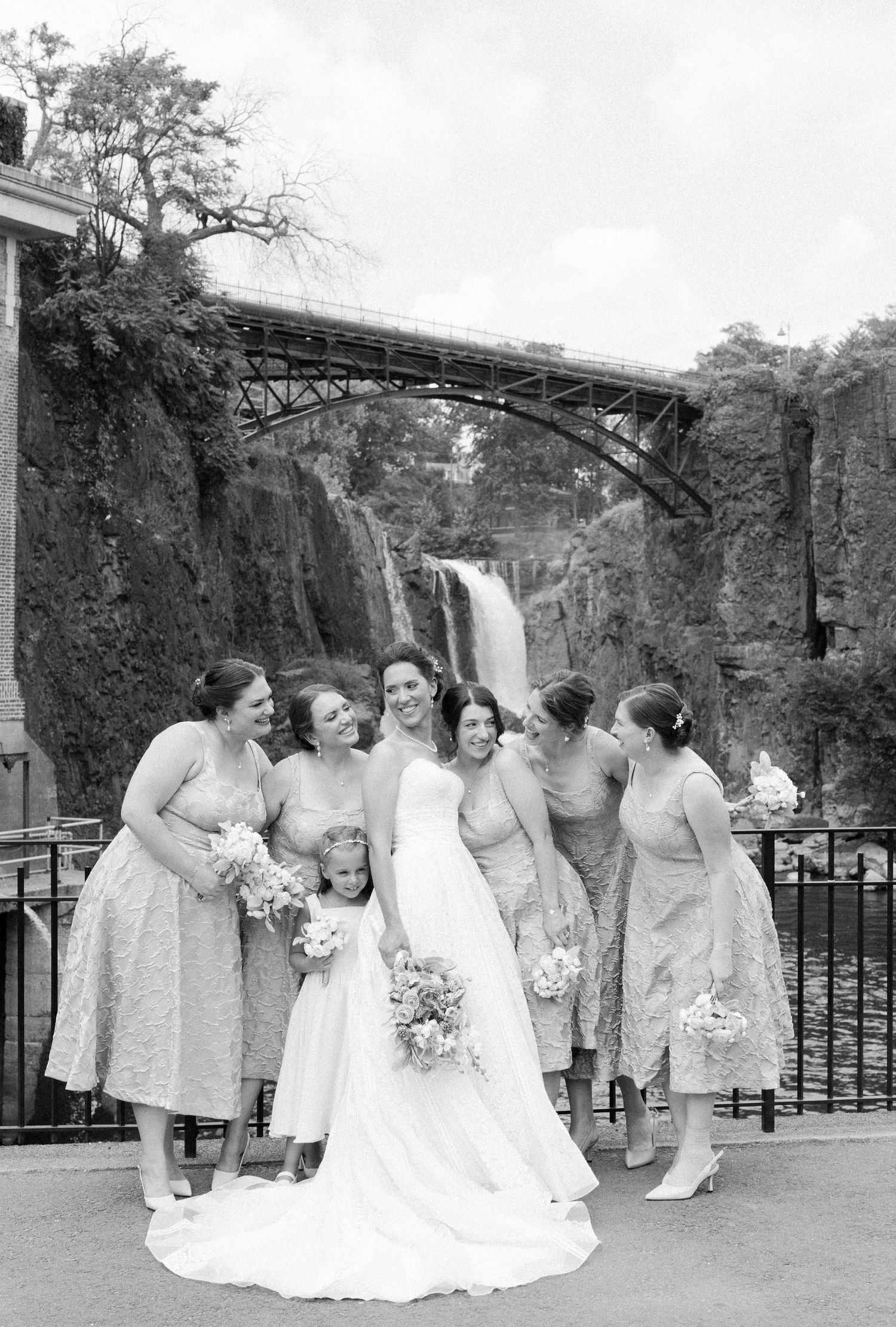 A vintage black and white wedding photo showing a bridal party gathered at Paterson Great Falls with an arched bridge above.