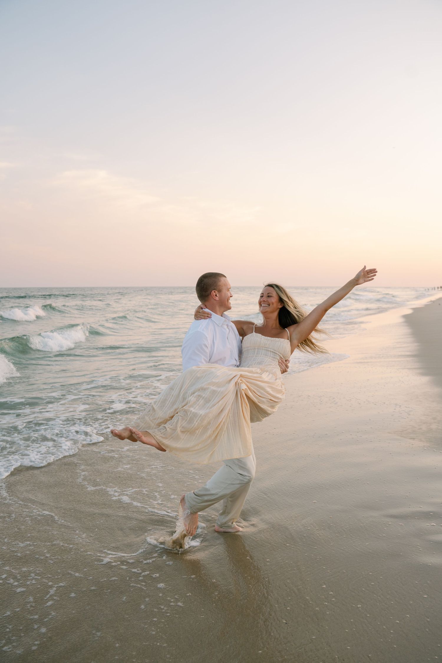 A couple dances playfully in the ocean waves at sunset, their white outfits glowing in the golden light.