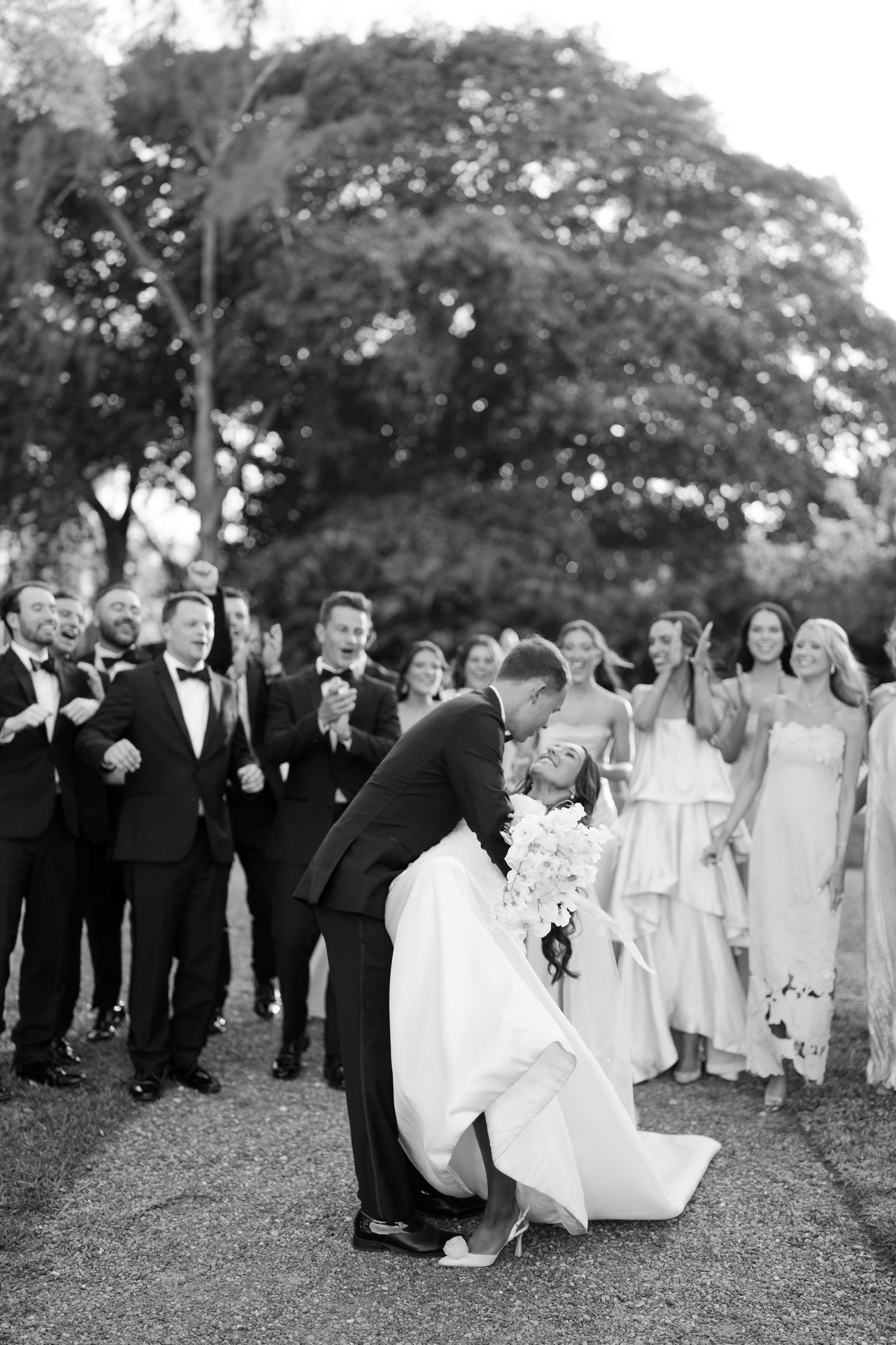 Black and white photo of groom dipping bride for a kiss surrounded by cheering wedding party.