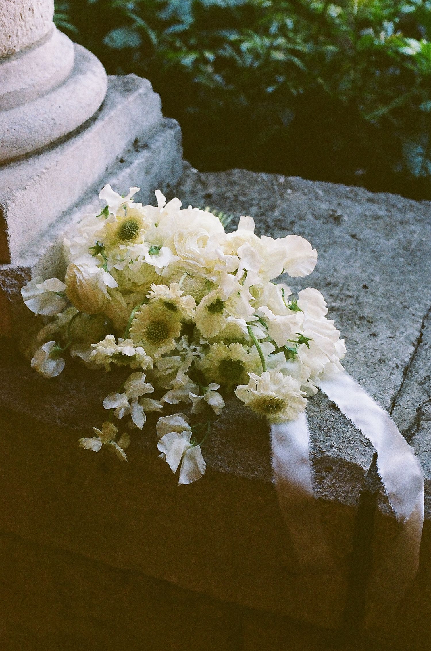 White wedding bouquet with roses and daisies tied with ribbon resting on stone steps outdoors.