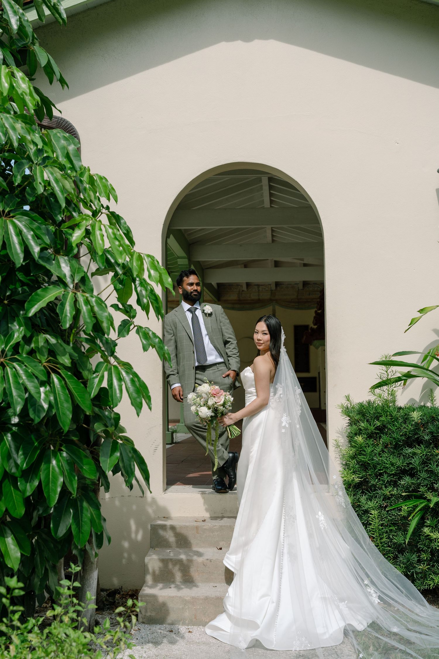 Bride in white gown and groom in gray suit pose in arched doorway surrounded by lush greenery at wedding venue.