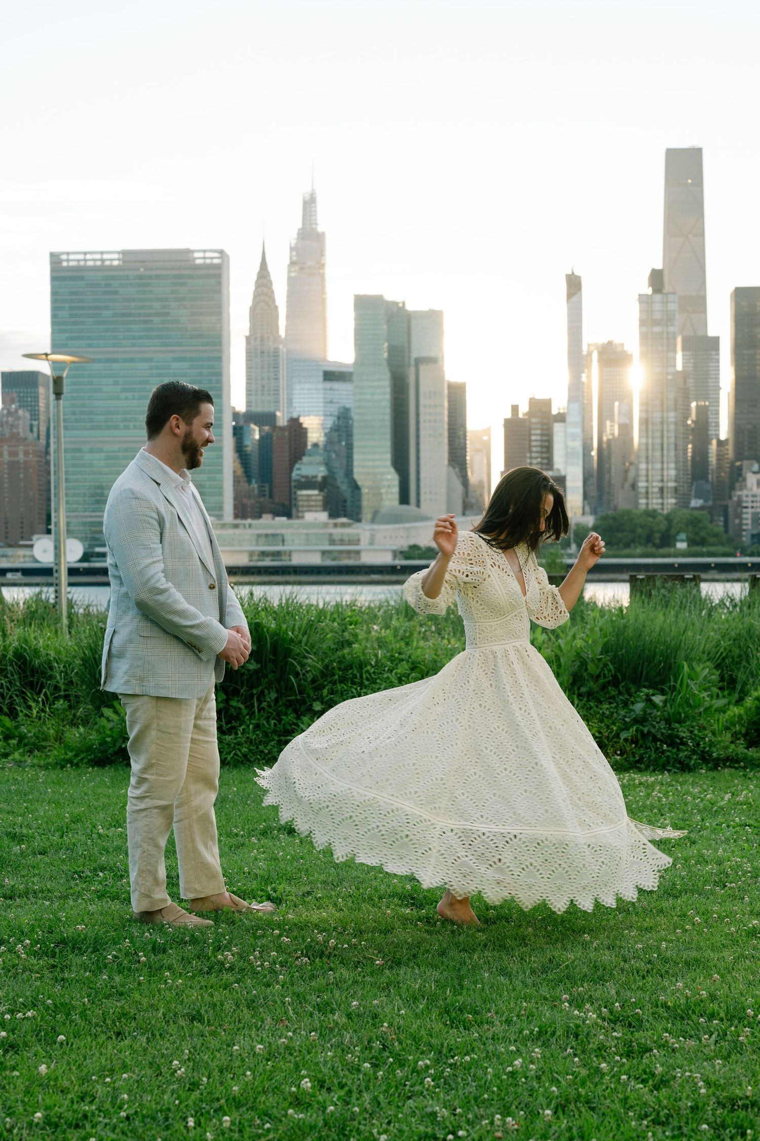 Couple dancing on grass with NYC skyline and golden sunset backdrop, woman in white eyelet dress twirling.