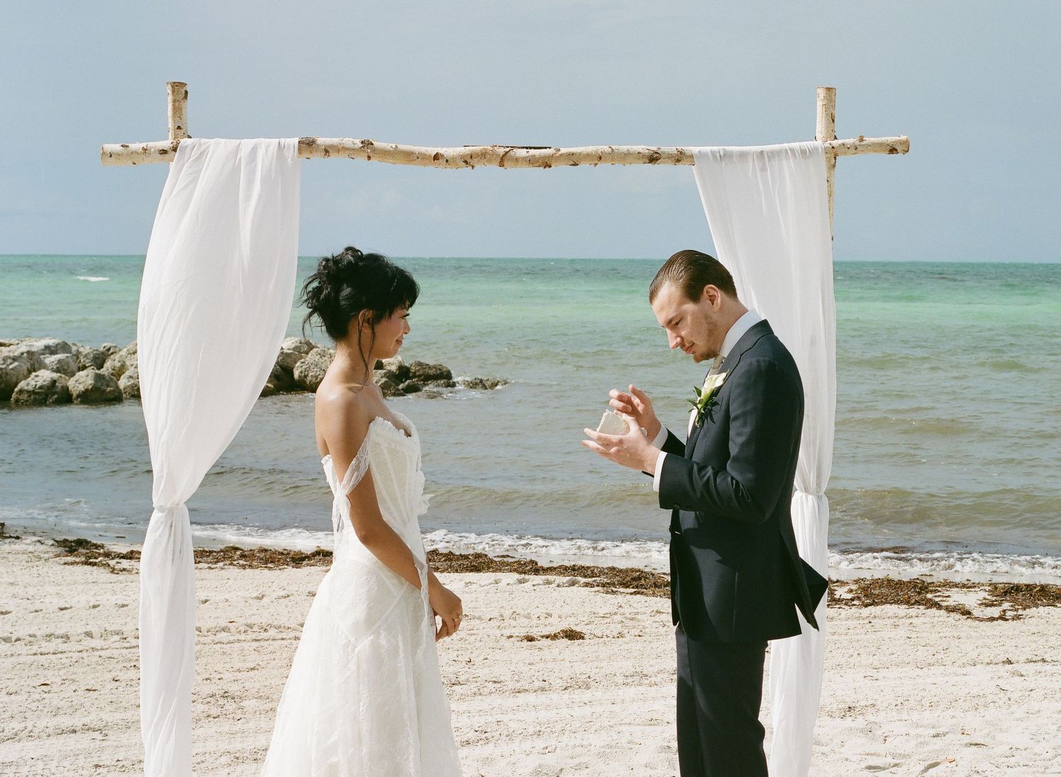 Couple exchanging vows at a romantic beach wedding ceremony under a white draped arch by the ocean.