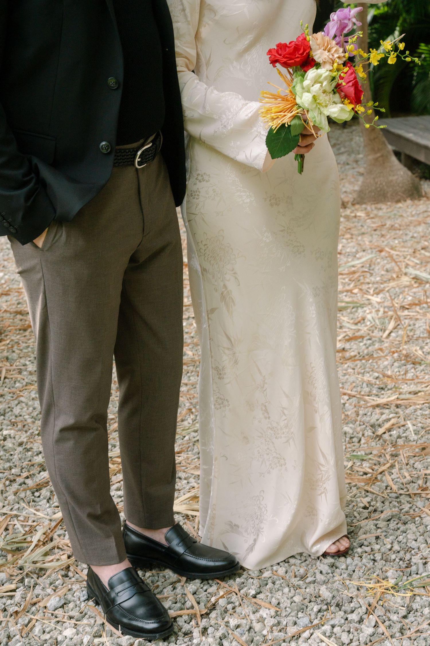 Close-up view of formal shoes and white wedding dress hem on gravel ground.