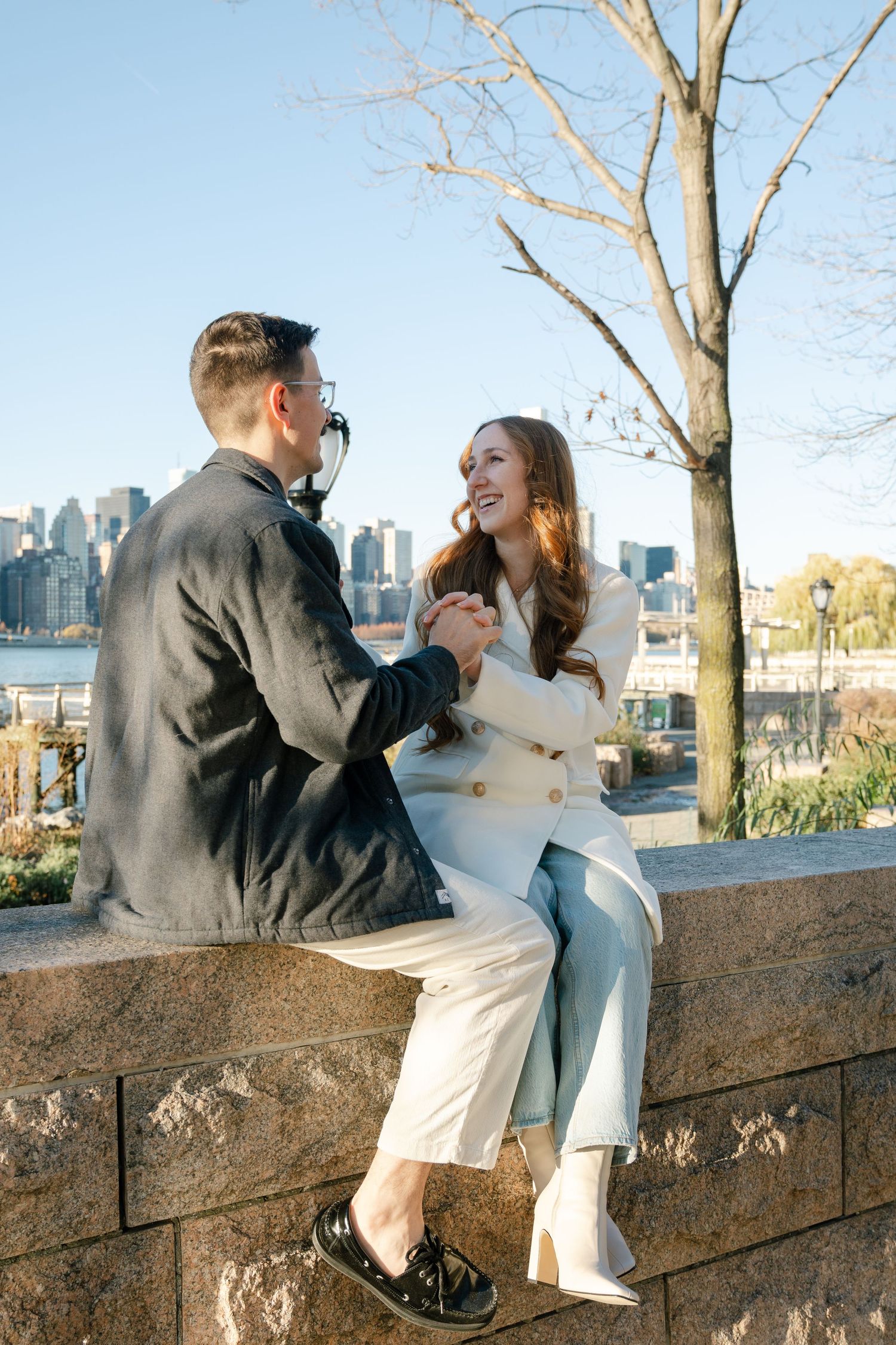 A romantic couple sits on a stone wall overlooking a city skyline during sunset.